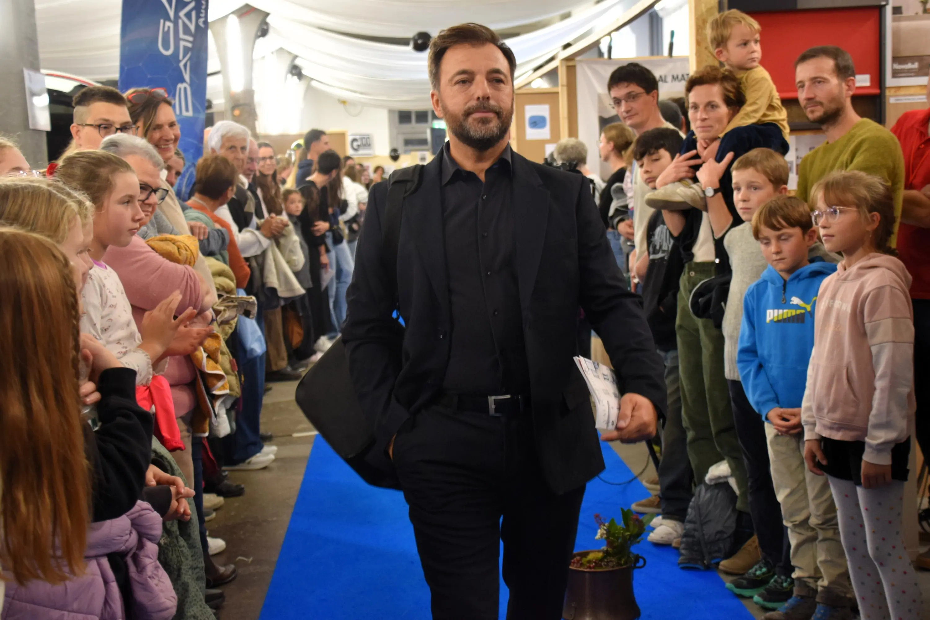 Un uomo in abito elegante cammina su un tappeto blu, circondato da una folla di persone che lo osservano. Alcuni bambini sono tra il pubblico, mentre altri adulti applaudono e sorridono. L'ambiente appare festoso e affollato.