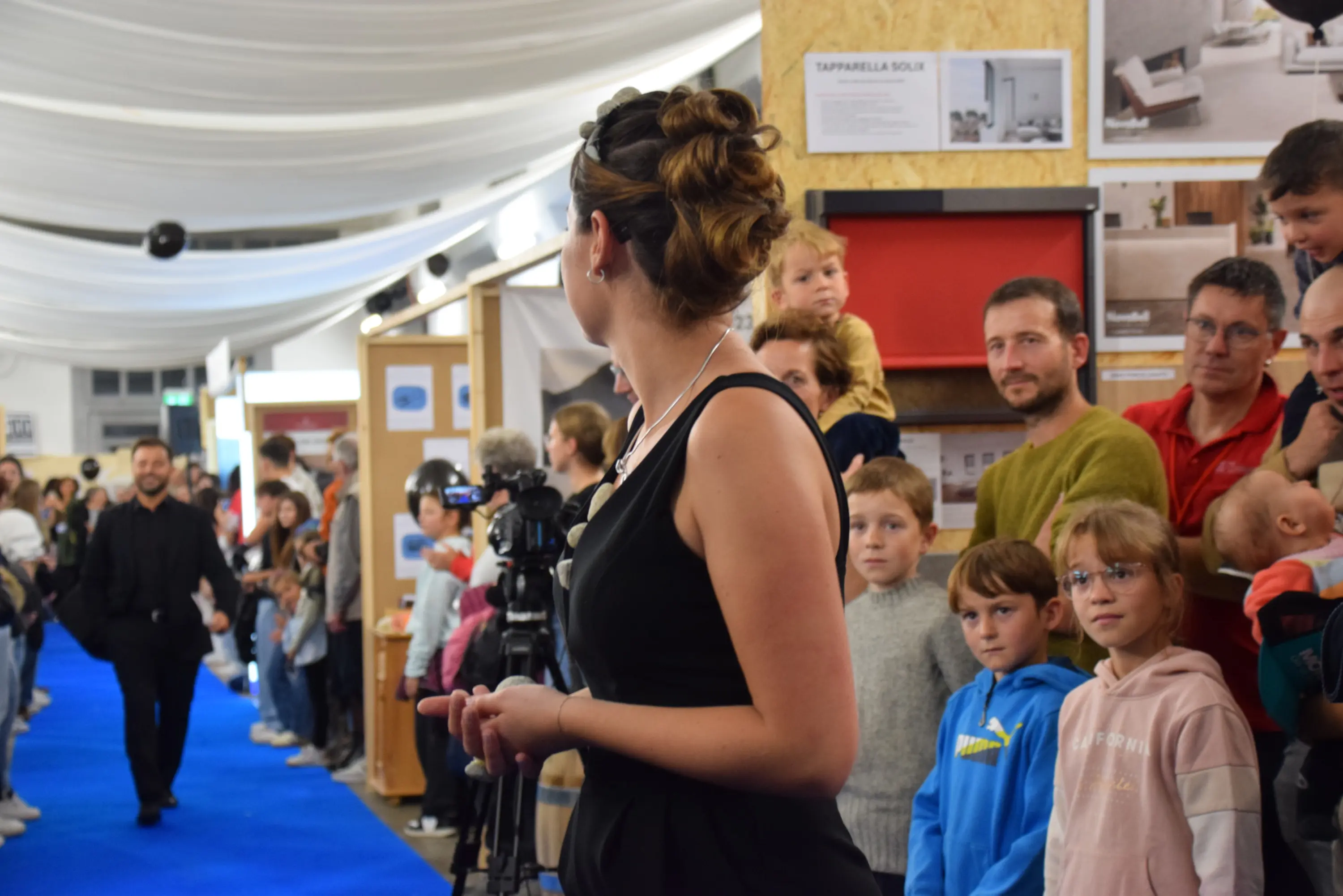 Una passerella con una modella in abito nero mentre un pubblico vario osserva. Al centro, un fotografo è in attesa di scattare, circondato da adulti e bambini interessati. Il pavimento è blu e ci sono espositori sullo sfondo.