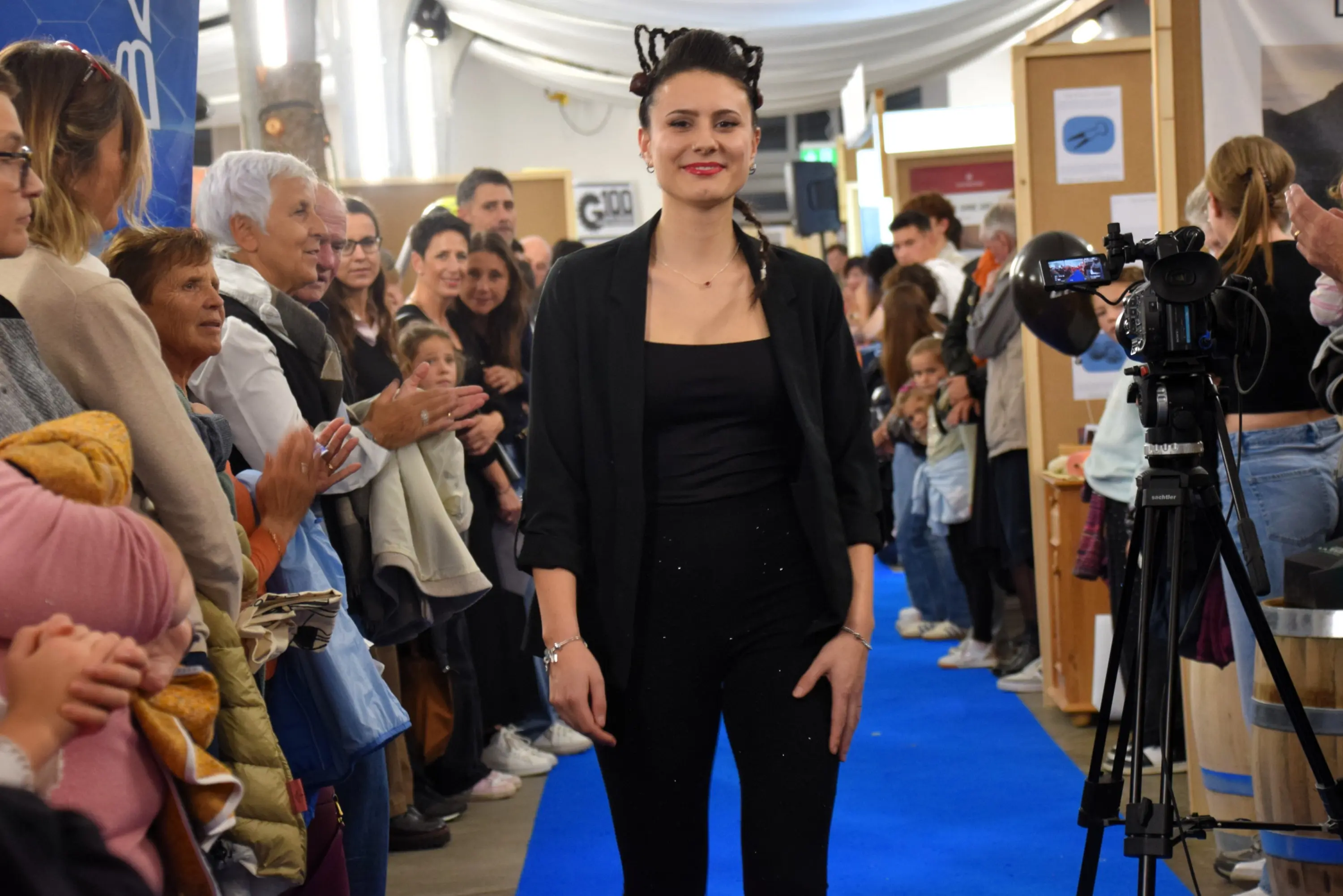 Un modello in abbigliamento elegante cammina su una passerella blu, circondato da un pubblico attento e incuriosito. La scena è vivace, con persone che applaudono e scattano foto.
