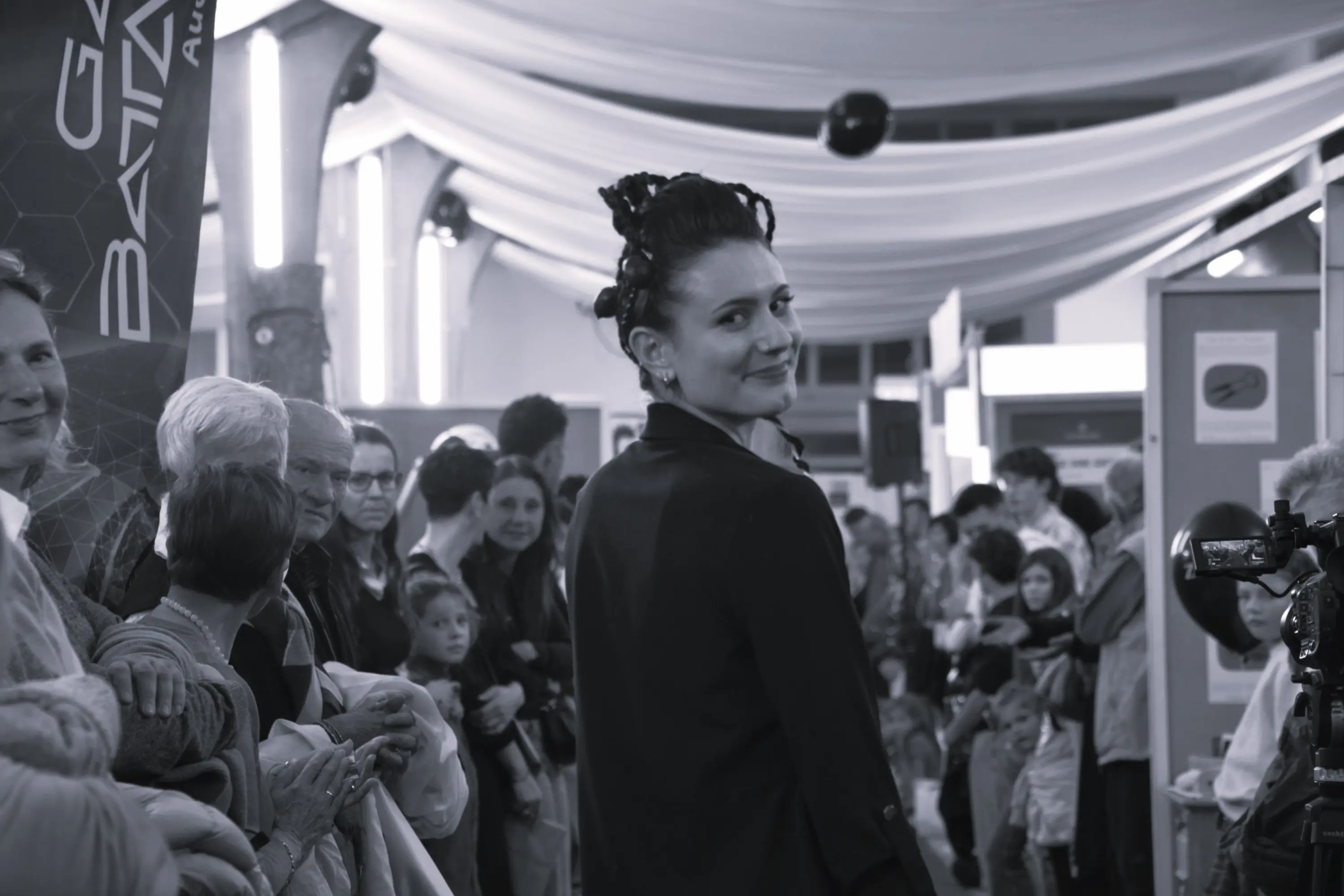 Una donna con capelli acconciati in nodi, indossa un abito elegante mentre sorride verso la telecamera. Intorno a lei si vede una folla di persone che osservano l'evento.