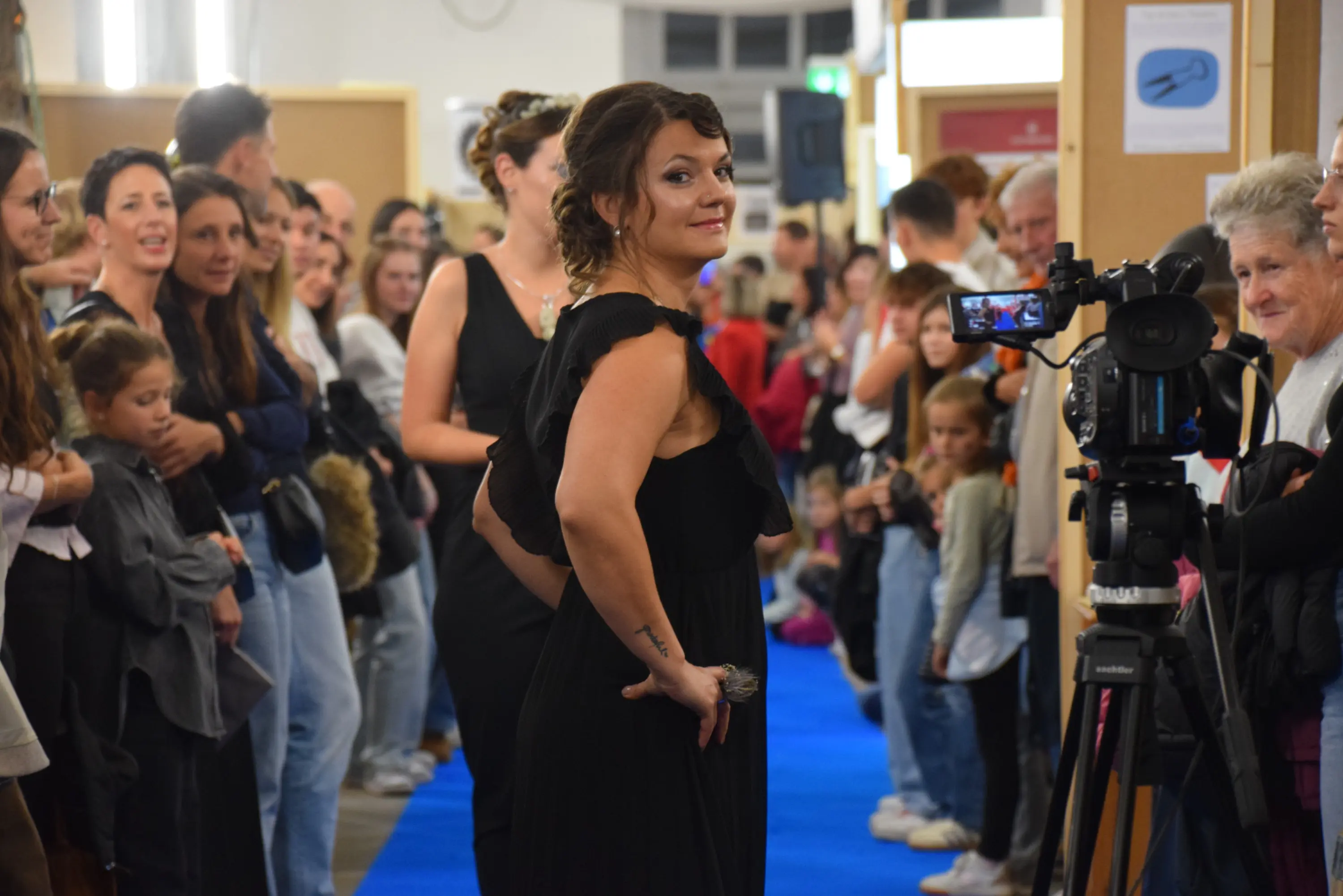 Una passerella affollata con un gruppo di persone che osservano, mentre una modella con un abito nero posiziona per le telecamere. Sullo sfondo, ci sono bambini e adulti che guardano l'evento.
