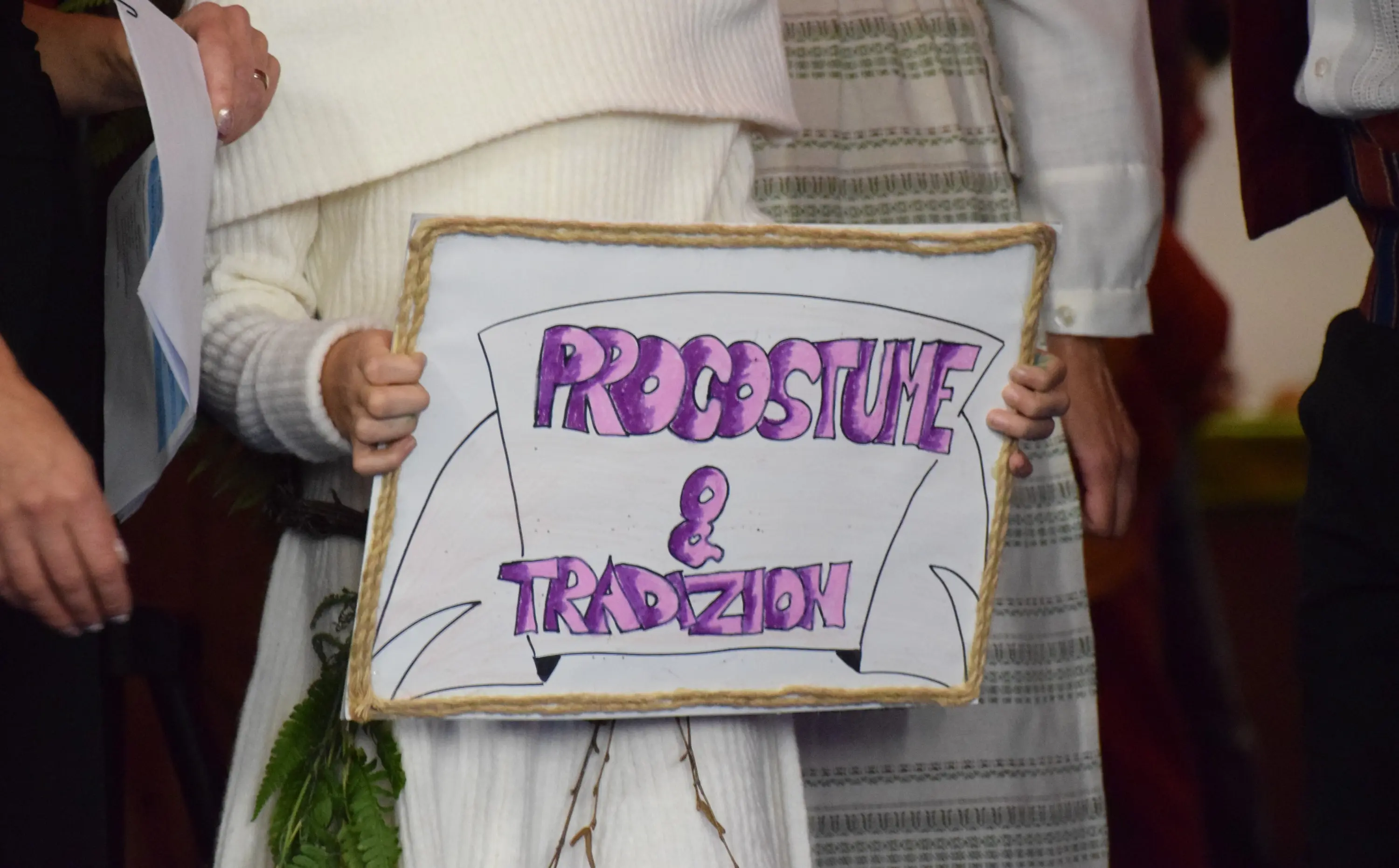 Un cartello con la scritta "Rocostume & Tradizion" decorato con colori vivaci. Una mano lo tiene mentre si svolge un evento.