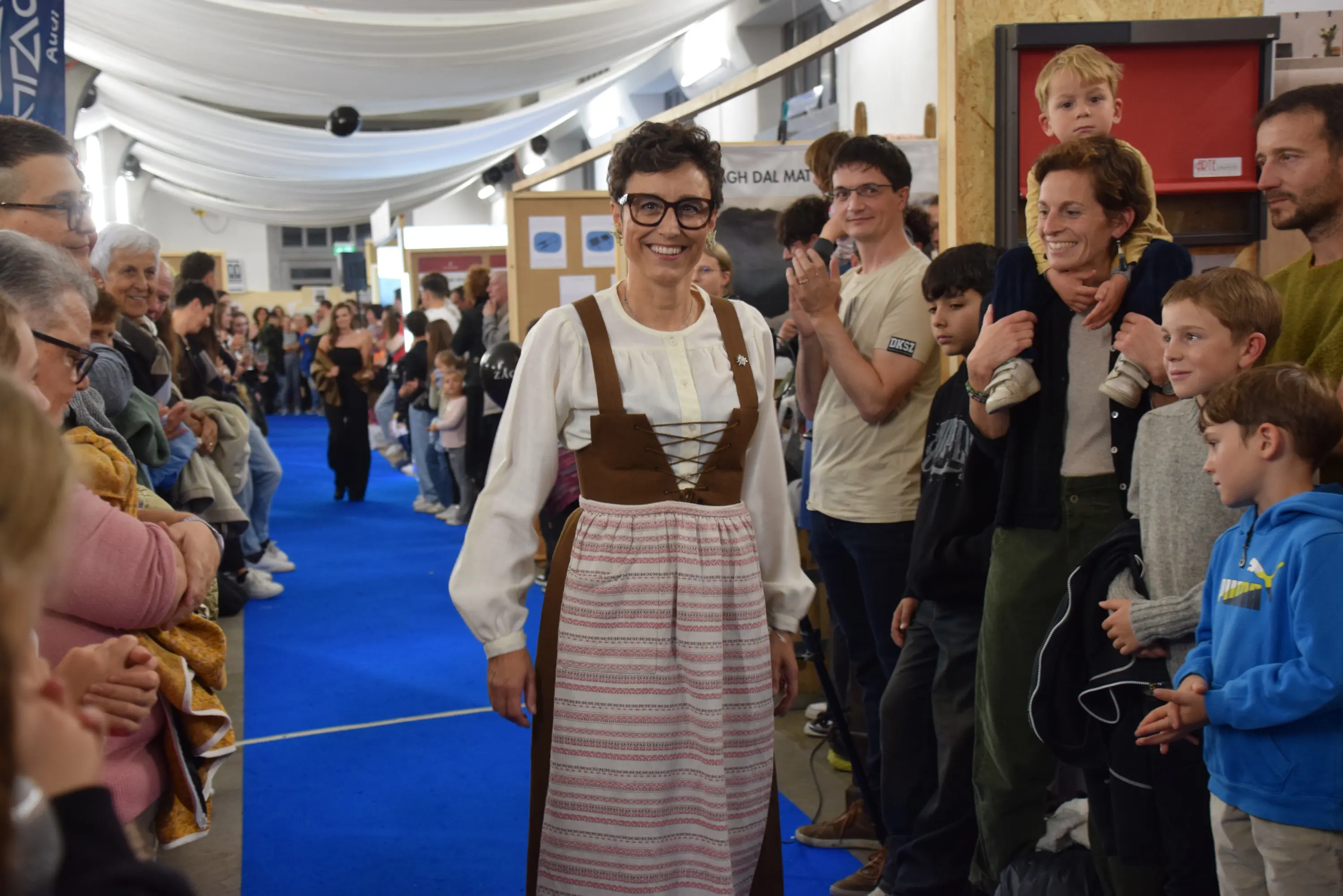 Donna che indossa un vestito tradizionale, sorridendo mentre cammina lungo un corridoio affollato. Persone applaudono e osservano attorno a lei, creando un'atmosfera festosa e vivace.