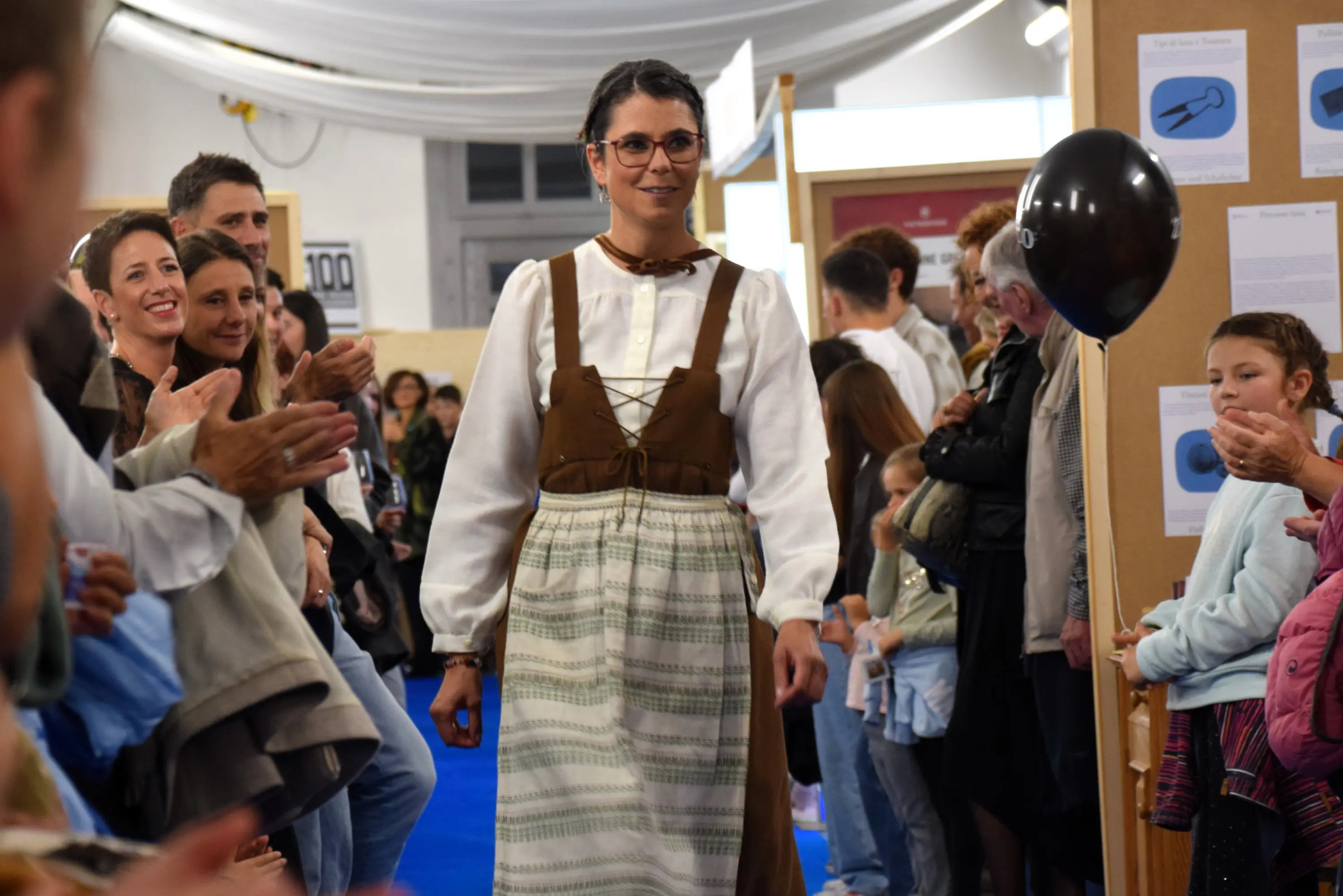 Una donna in abito tradizionale con occhiali passeggia lungo una passerella, mentre il pubblico applaude. Sullo sfondo si vedono espositori e decorazioni.