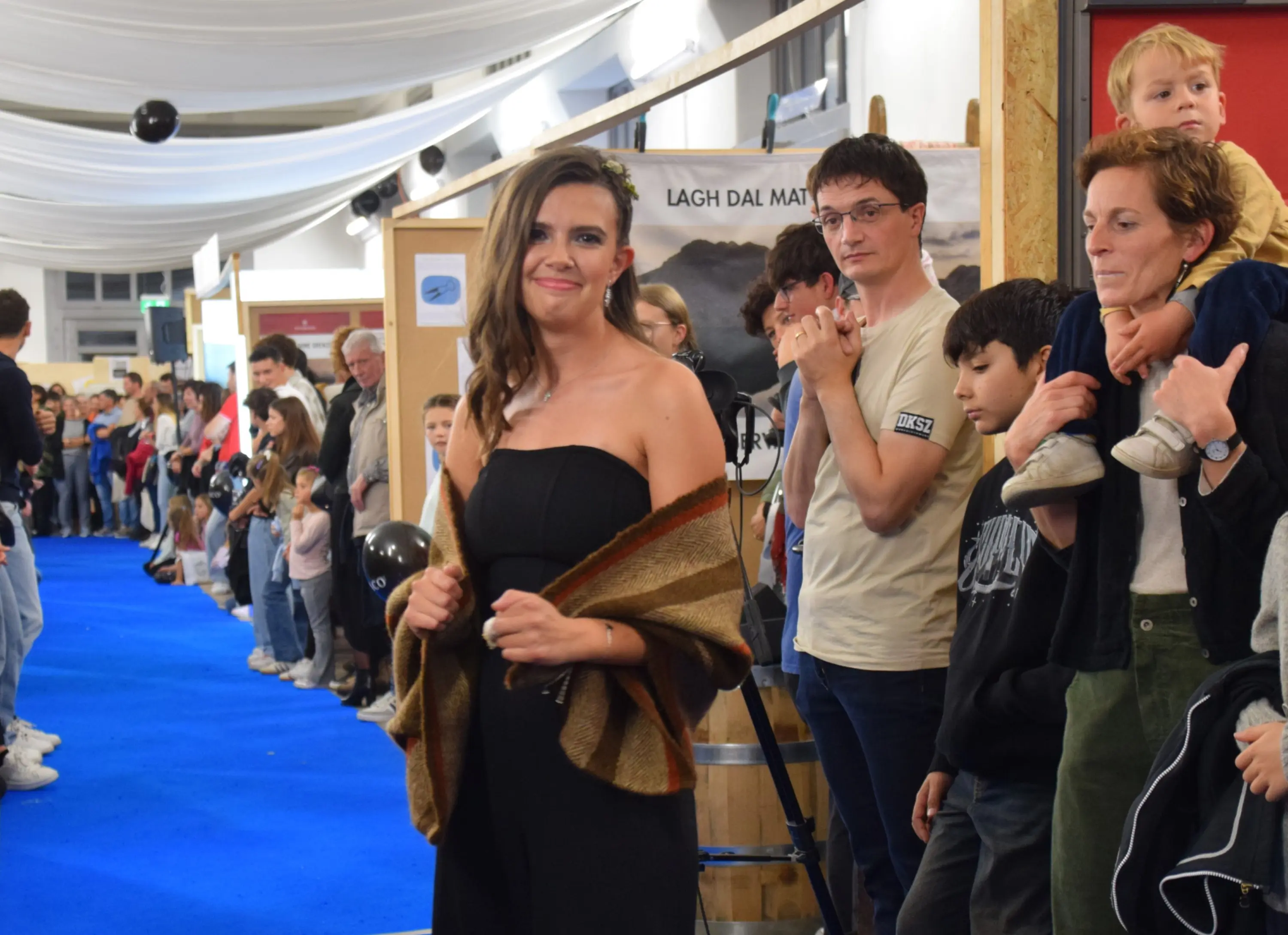 Una donna sorridente indossa un vestito nero e una sciarpa colorata, mentre si trova su un tappeto blu. Dietro di lei, una folla di persone di diverse età guarda con interesse.