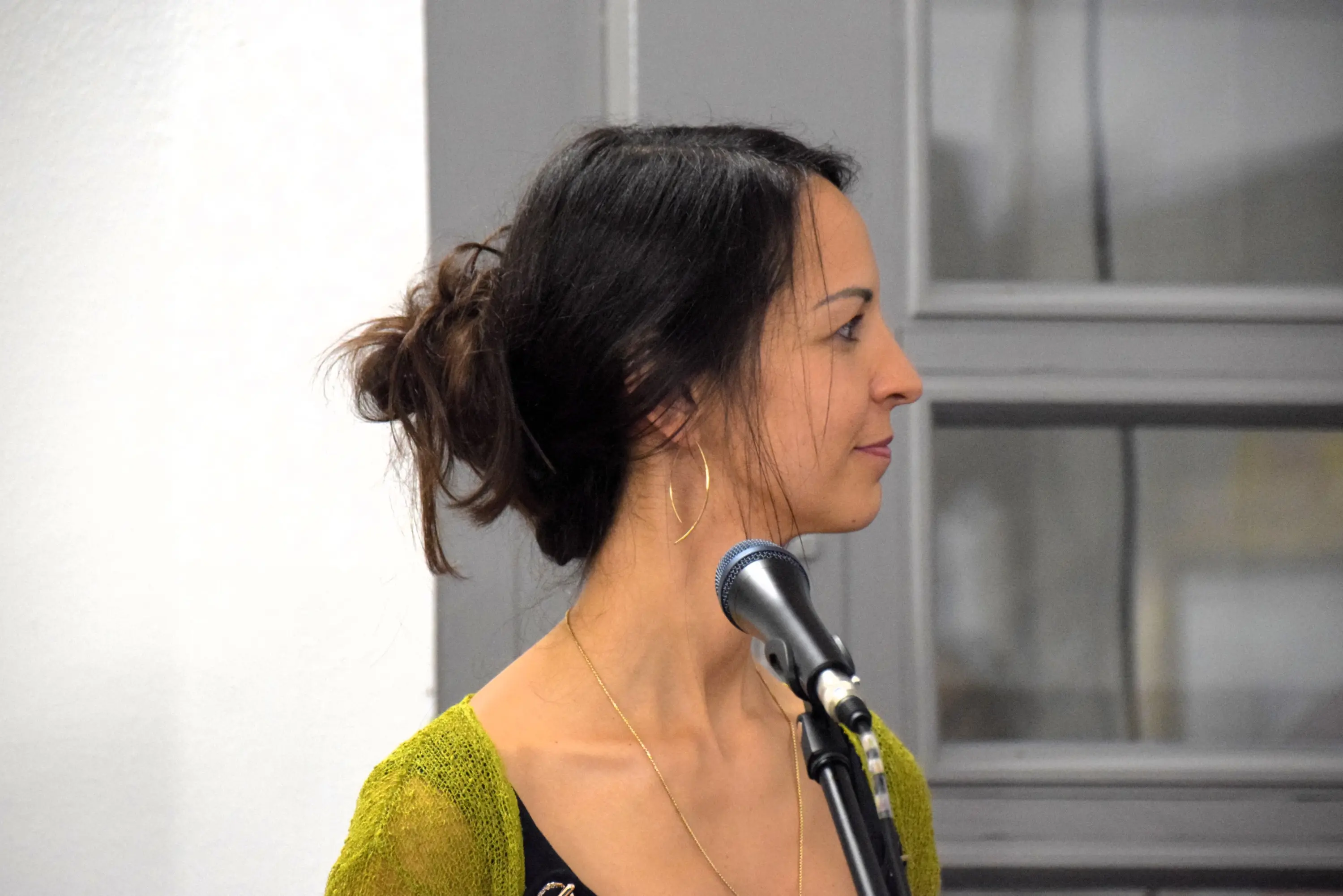Donna con capelli raccolti in uno chignon, abbigliamento scuro e cardigan verde. È di profilo e guarda verso sinistra, con un microfono davanti a sé. Le orecchie hanno orecchini a cerchio. Sullo sfondo si intravede una fines