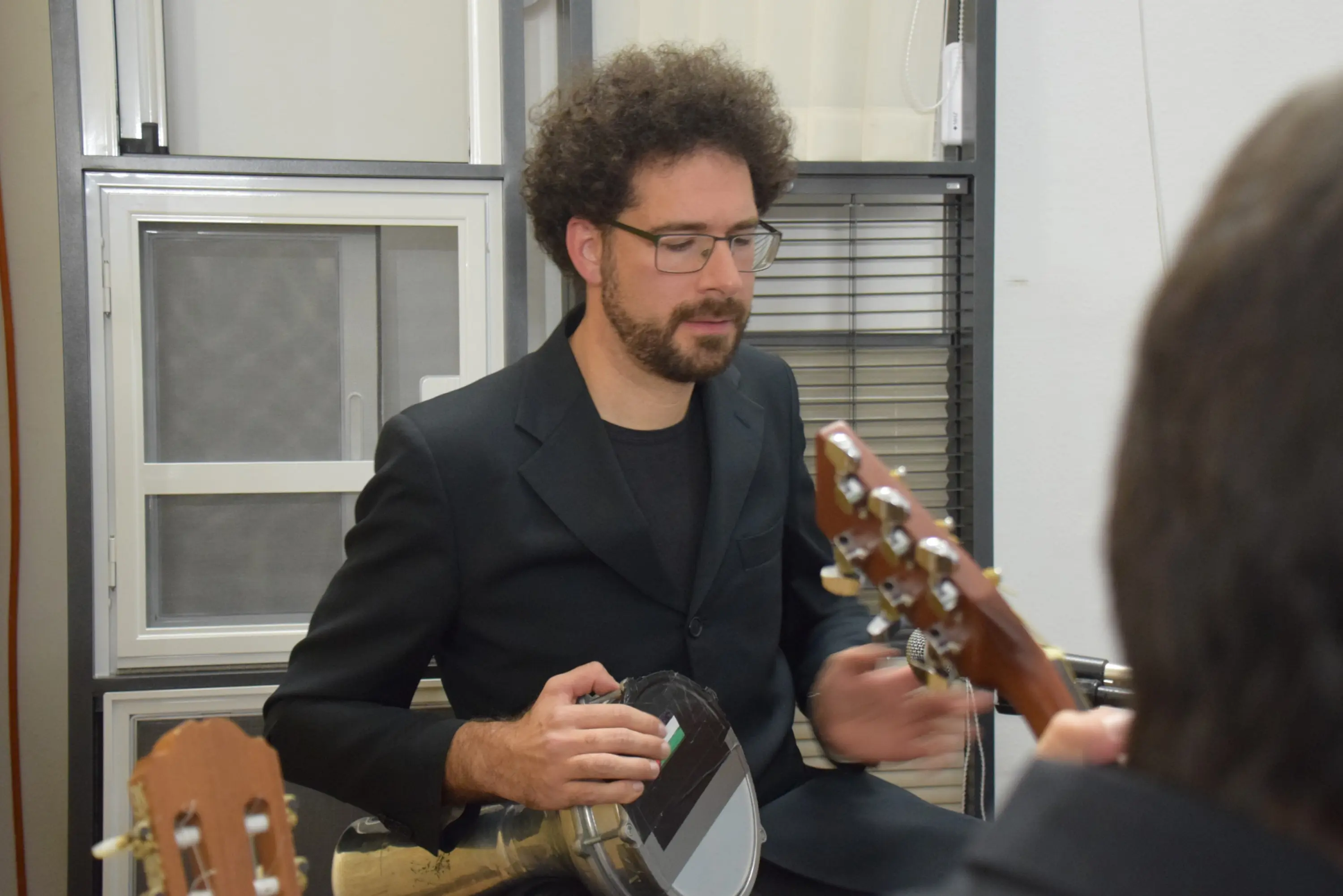 Uomo con occhiali e capelli ricci, vestito di nero, suona un banjo mentre si trova in un ambiente interno. Sullo sfondo si vedono strumenti musicali e finestre.