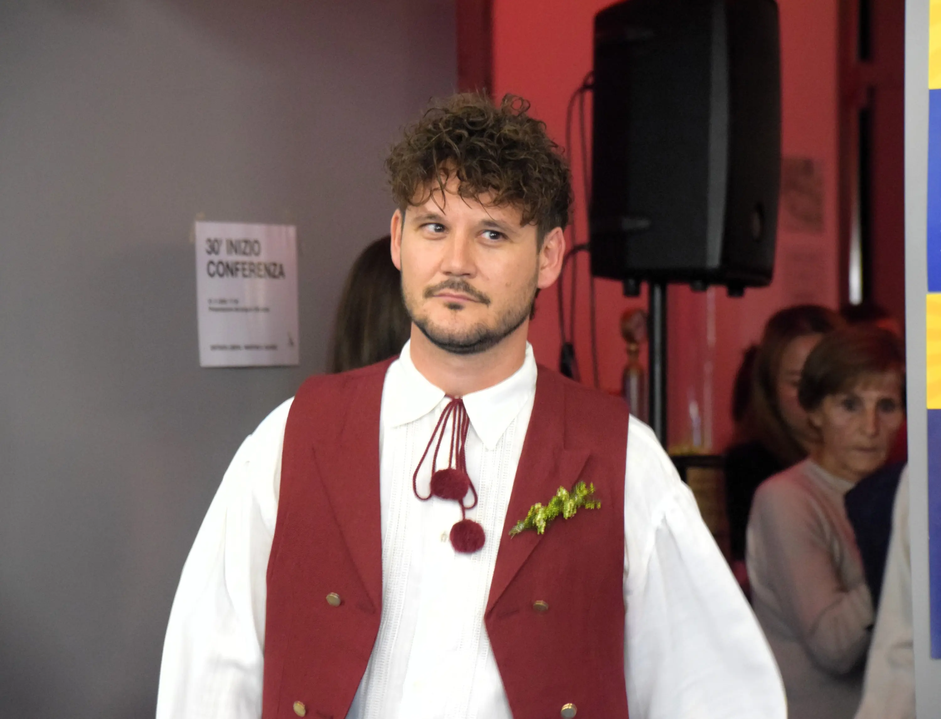 Uomo con capelli ricci e barba, indossa un abito tradizionale con giacca rossa e camicia bianca. Sul petto ha una decorazione verde. Sullo sfondo, si intravedono delle persone e un banner con scritte.