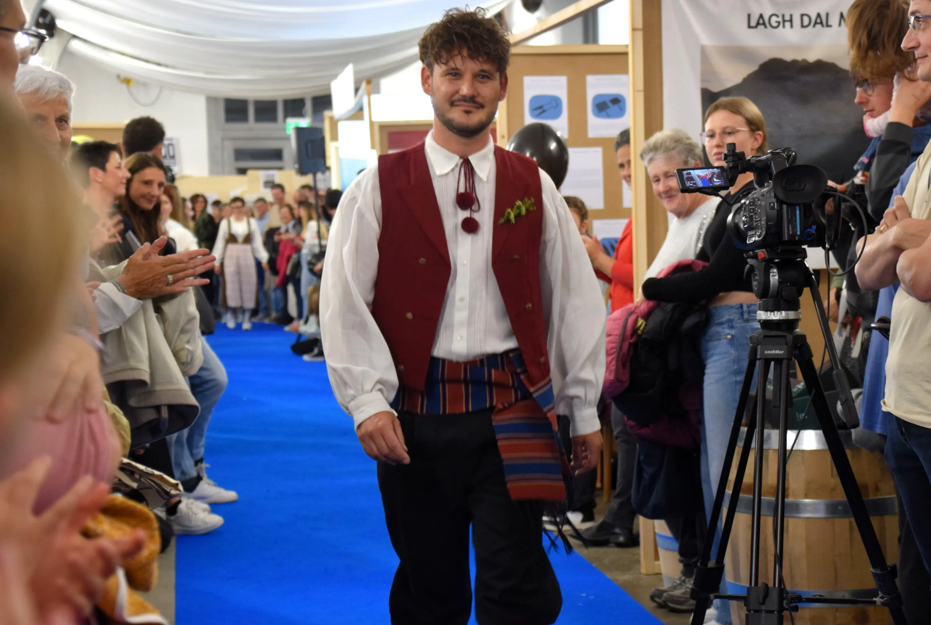 Un uomo in abiti tradizionali cammina su una passerella blu, circondato da un pubblico che applaude. Sullo sfondo, si vedono bancarelle e una telecamera che registra.