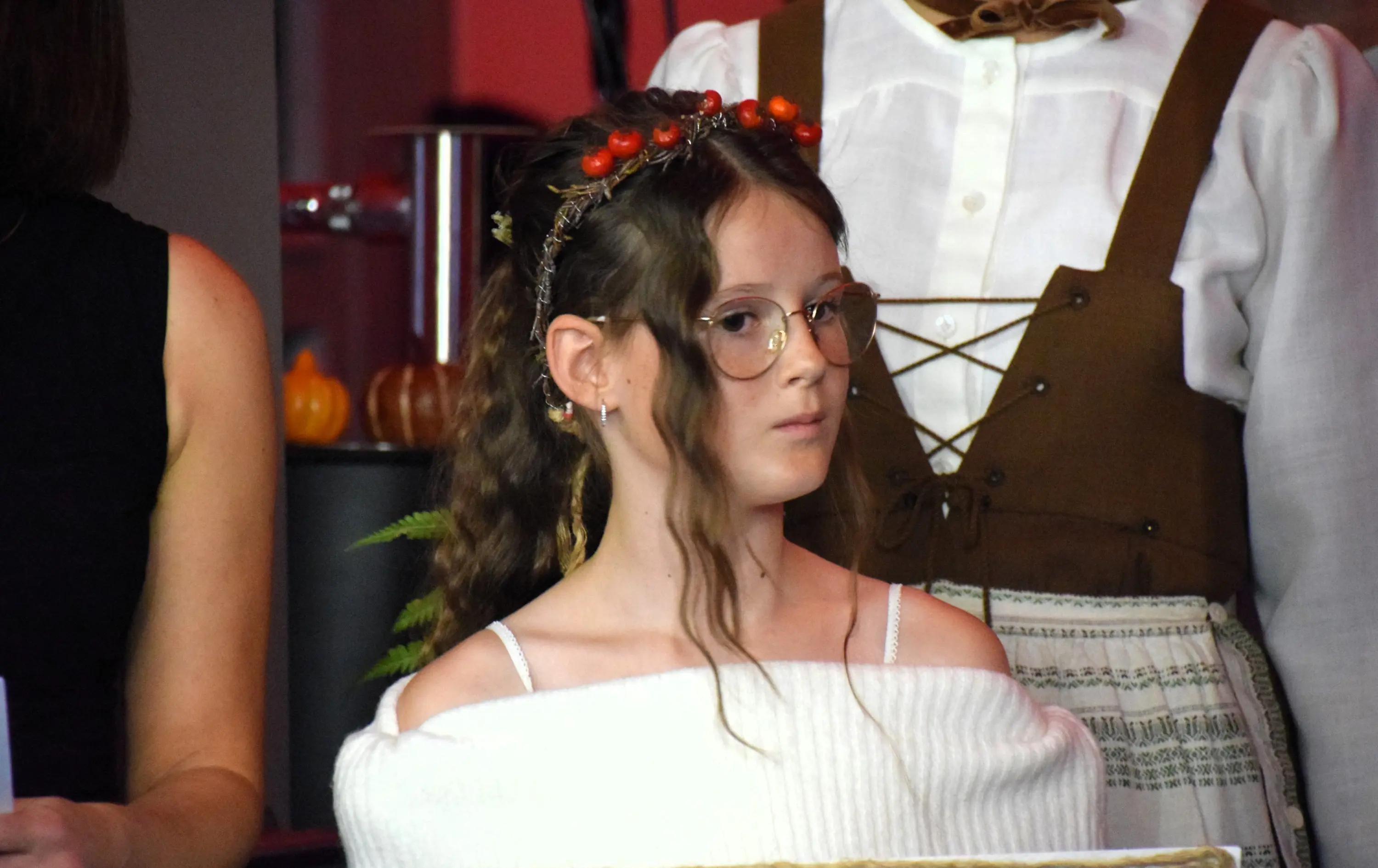 Ragazza con occhiali e corona di fiori rossi, indossa un maglione bianco a spalle scoperte e guarda in modo pensieroso. Nel sfondo, un'altra persona in abiti tradizionali.