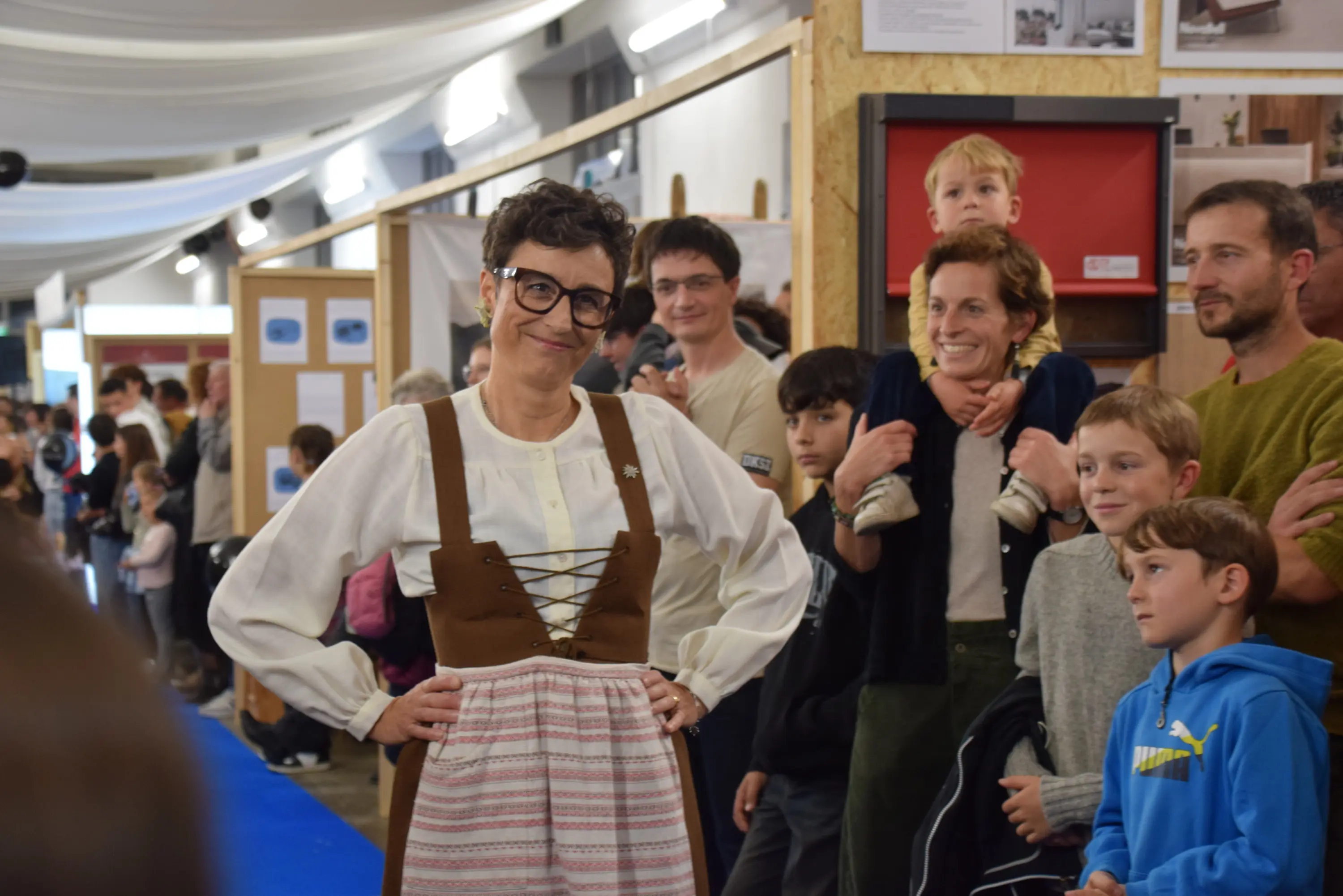 Una donna sorridente in abiti tradizionali tirolesi posa davanti a un gruppo di persone di diverse età, tra cui bambini. Dietro di lei, un ambiente di esposizione con pannelli informativi.