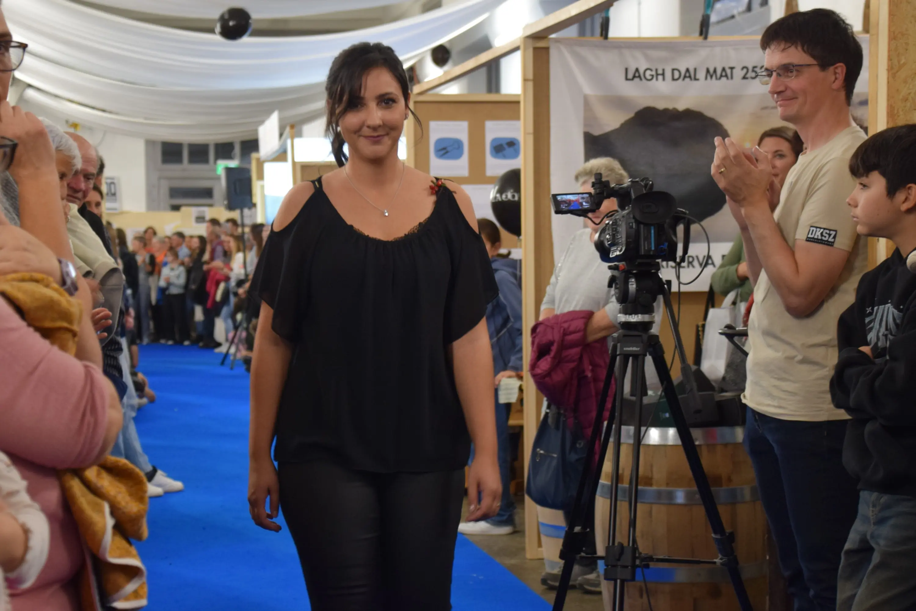 Una donna in abbigliamento elegante cammina lungo una passerella, mentre il pubblico la osserva. Sullo sfondo, una telecamera riprende l'evento e si vedono alcune persone che applaudono.