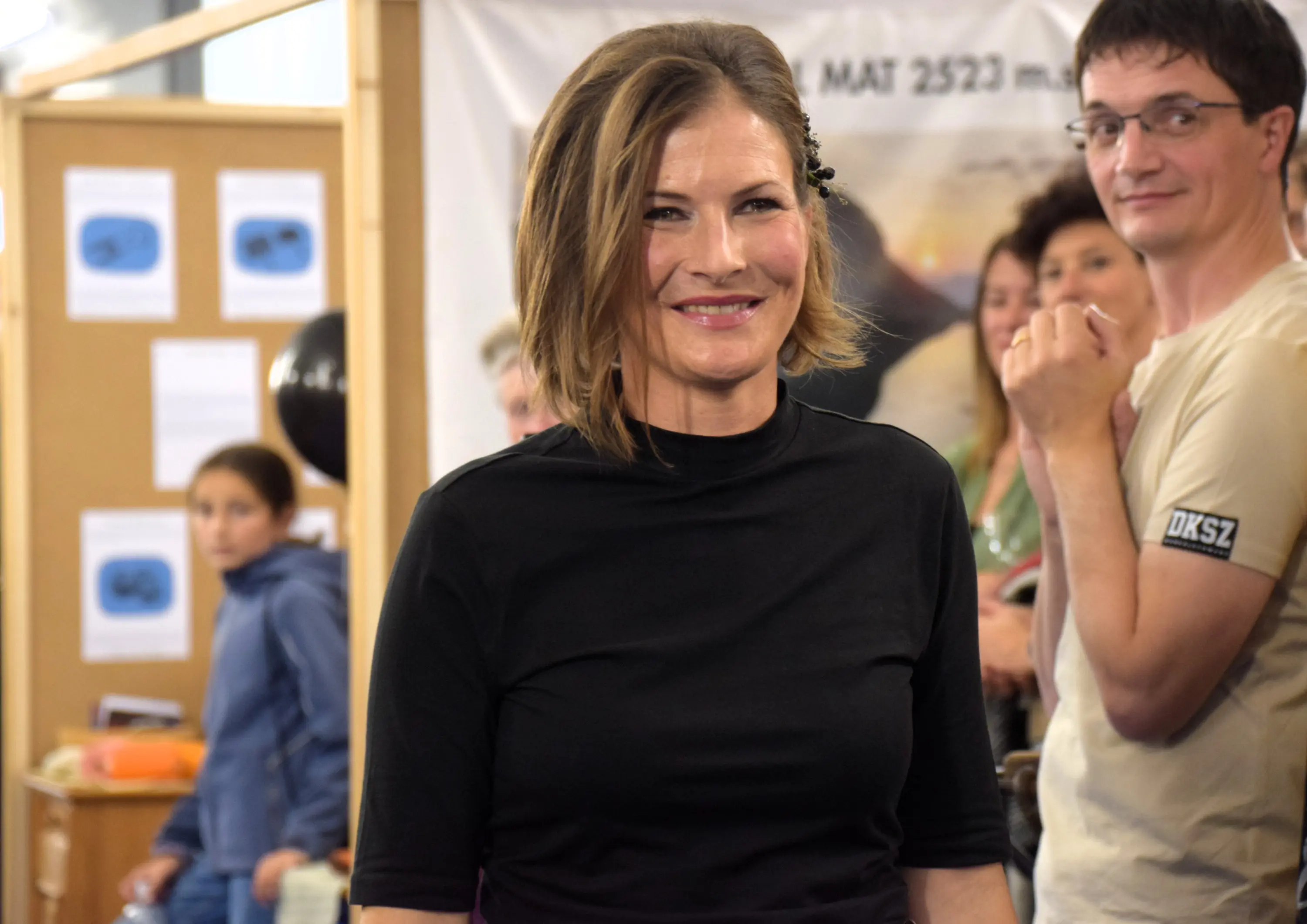 Una donna sorride indossando una maglietta nera, mentre si trova in una folla. Sullo sfondo, ci sono persone in un ambiente probabilmente decorato per un evento.