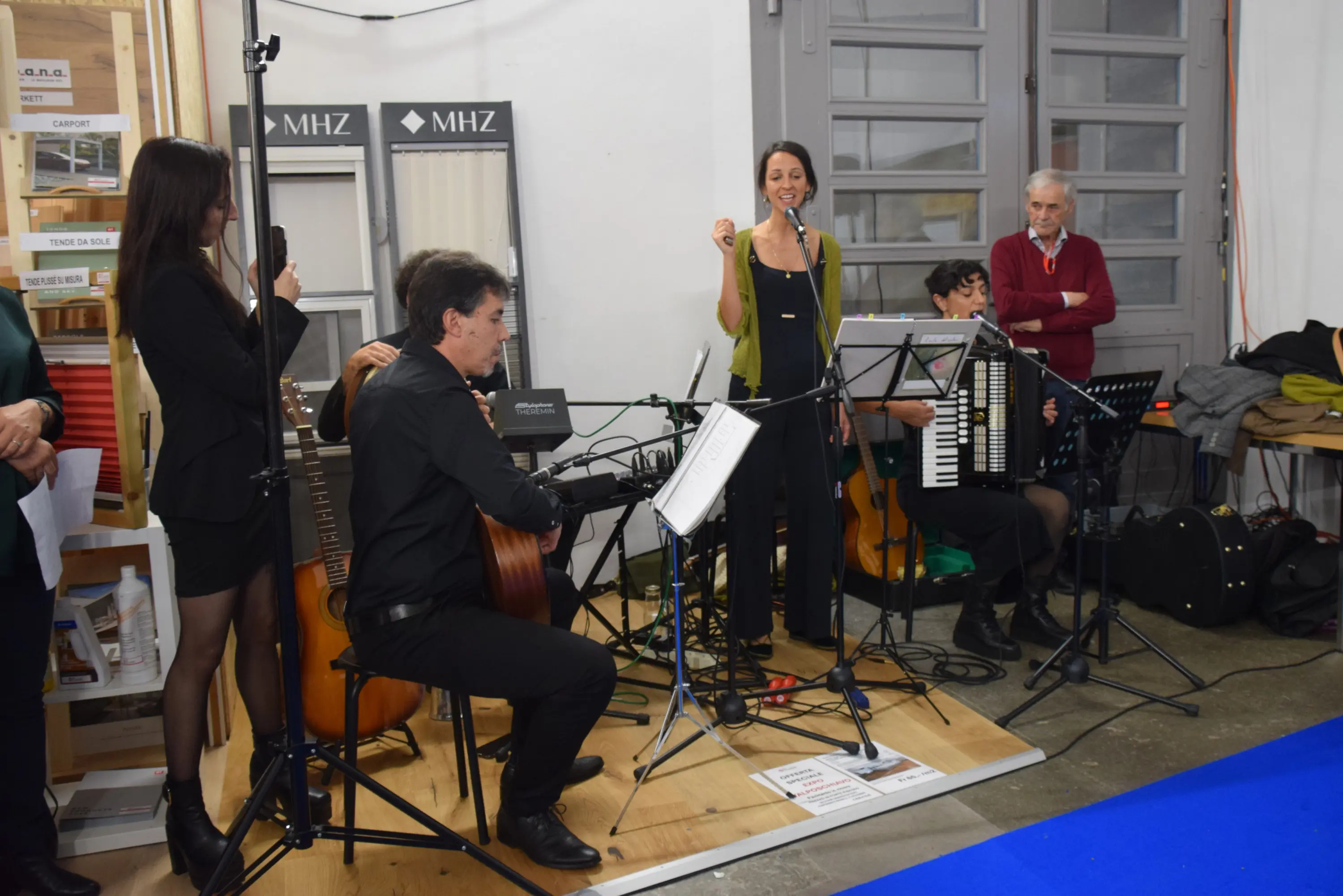 Un gruppo musicale composto da cinque musicisti che suonano strumenti come chitarra, fisarmonica e violino. Una cantante si esibisce al centro, mentre gli altri membri assistono. L'ambientazione sembra essere un evento o una manifestazione in un locale.