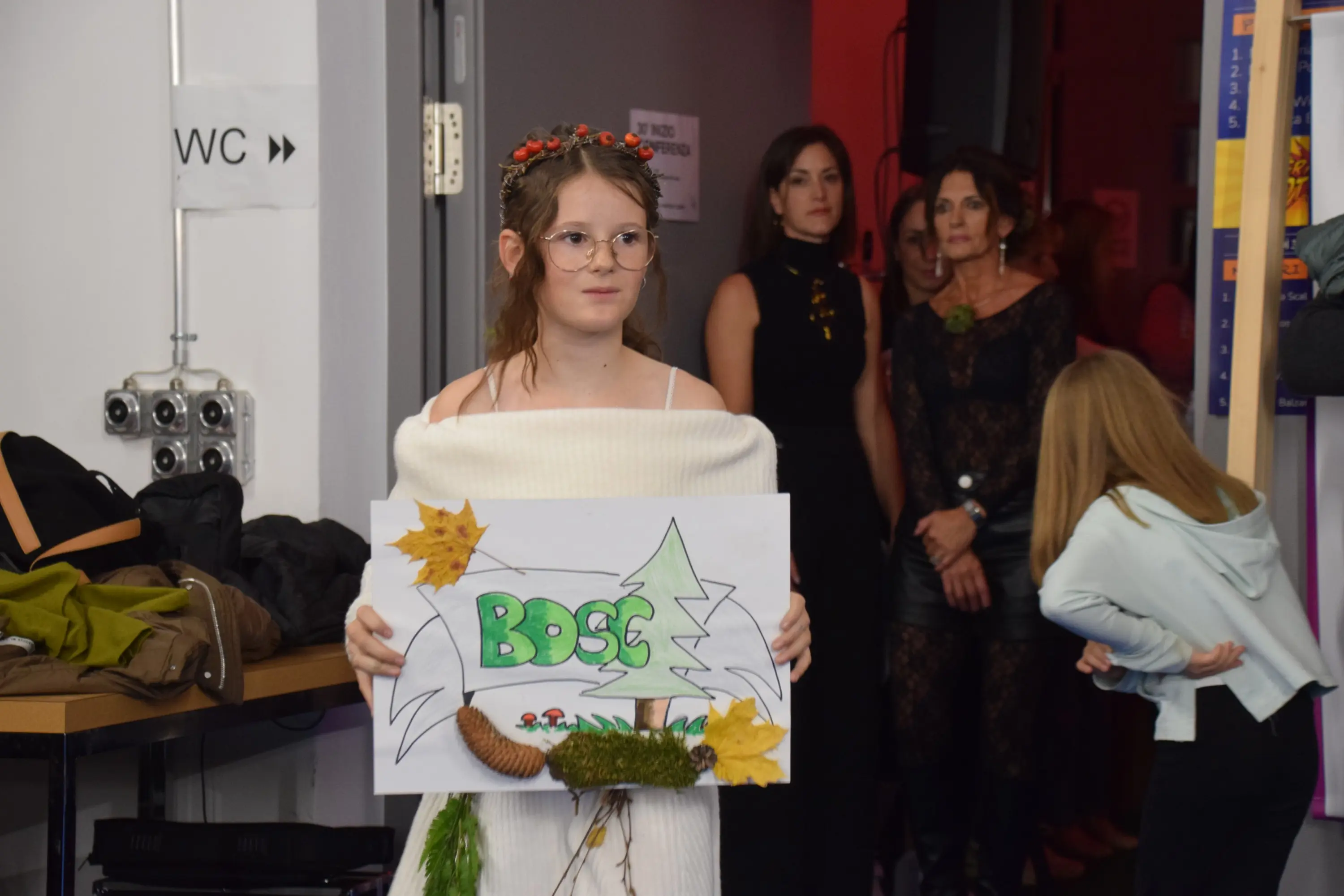 Ragazza con occhiali e capelli raccolti, indossa un abito bianco e tiene in mano un cartellone decorato con foglie e disegni di alberi. Sullo sfondo, alcune persone osservano l’evento.