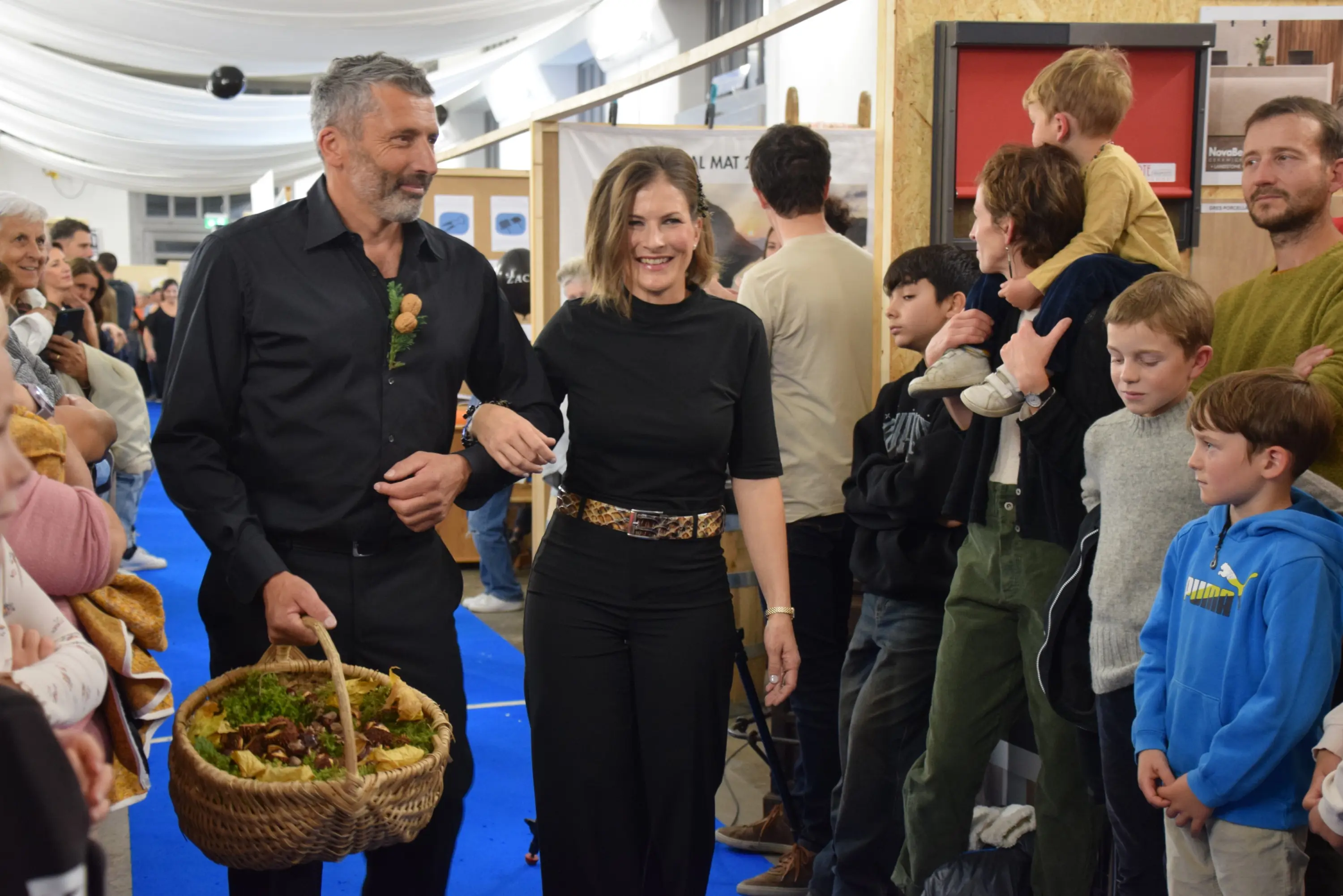 Una coppia sorridente cammina insieme, il uomo porta un cesto pieno di cibo. Attorno a loro, un gruppo di persone, tra cui bambini curiosi, osserva con interesse. L'ambiente è vivace e festoso.