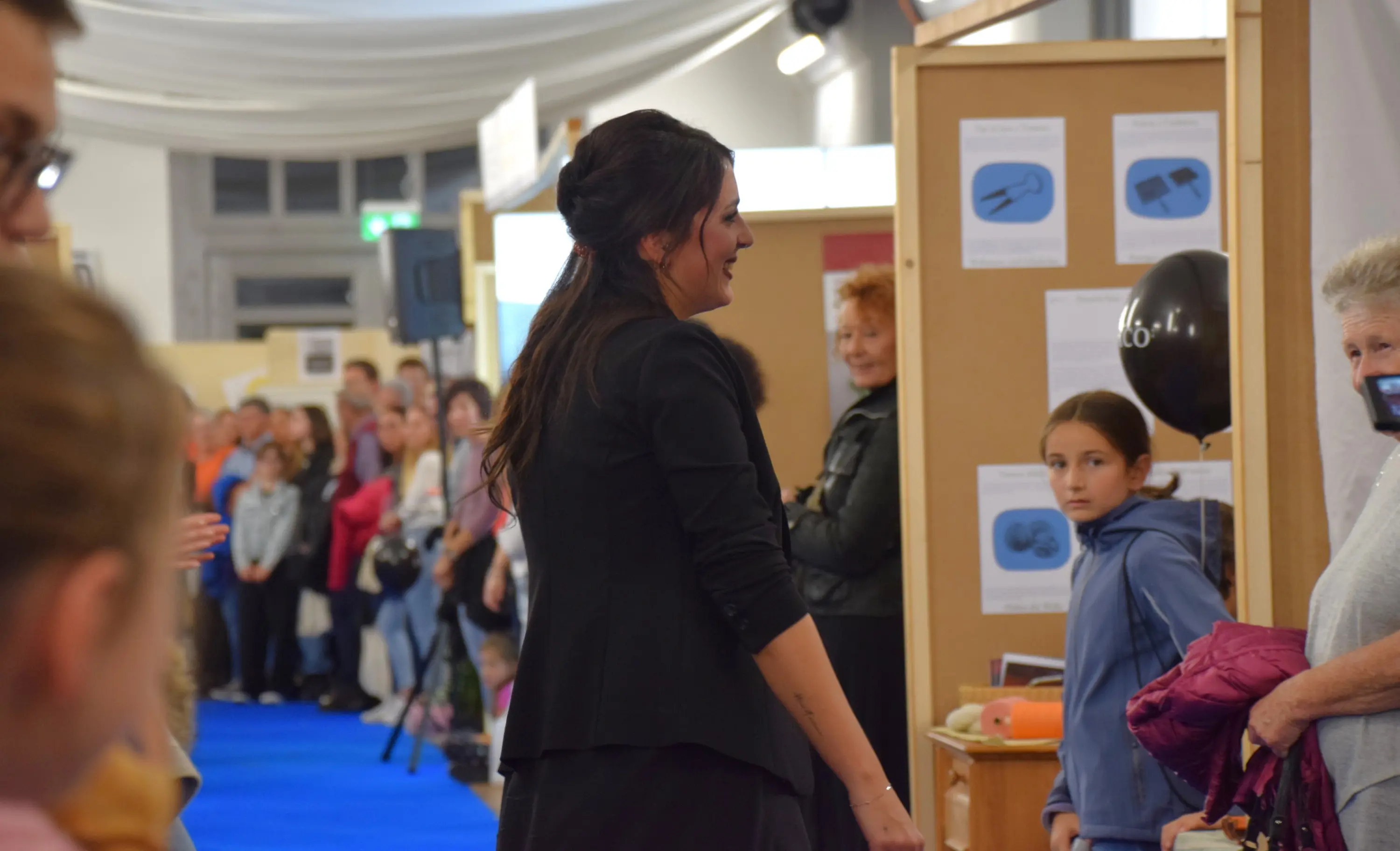 Una donna con i capelli scuri indossa un abito elegante mentre cammina verso un pubblico numeroso. Alcuni spettatori osservano con interesse, mentre una bambina e un'altra donna sorridono in primo piano. La scena si svolge in un ambiente espositivo con pannelli inform