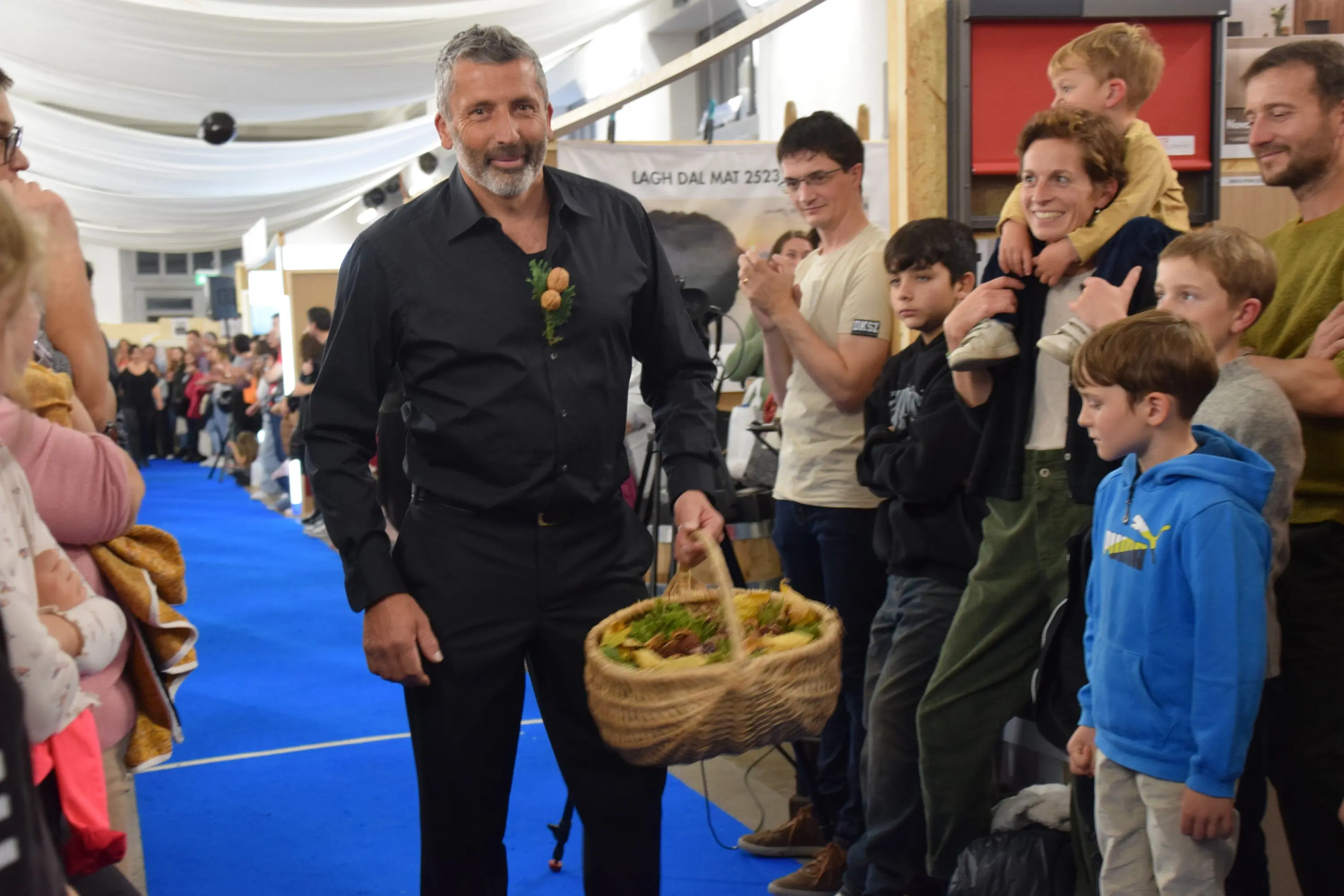 Un uomo con una camicia nera e un cestino pieno di frutta cammina lungo una passerella blu, circondato da un pubblico entusiasta. Alcune persone applaudono e sorridono, mentre i bambini osservano con interesse.