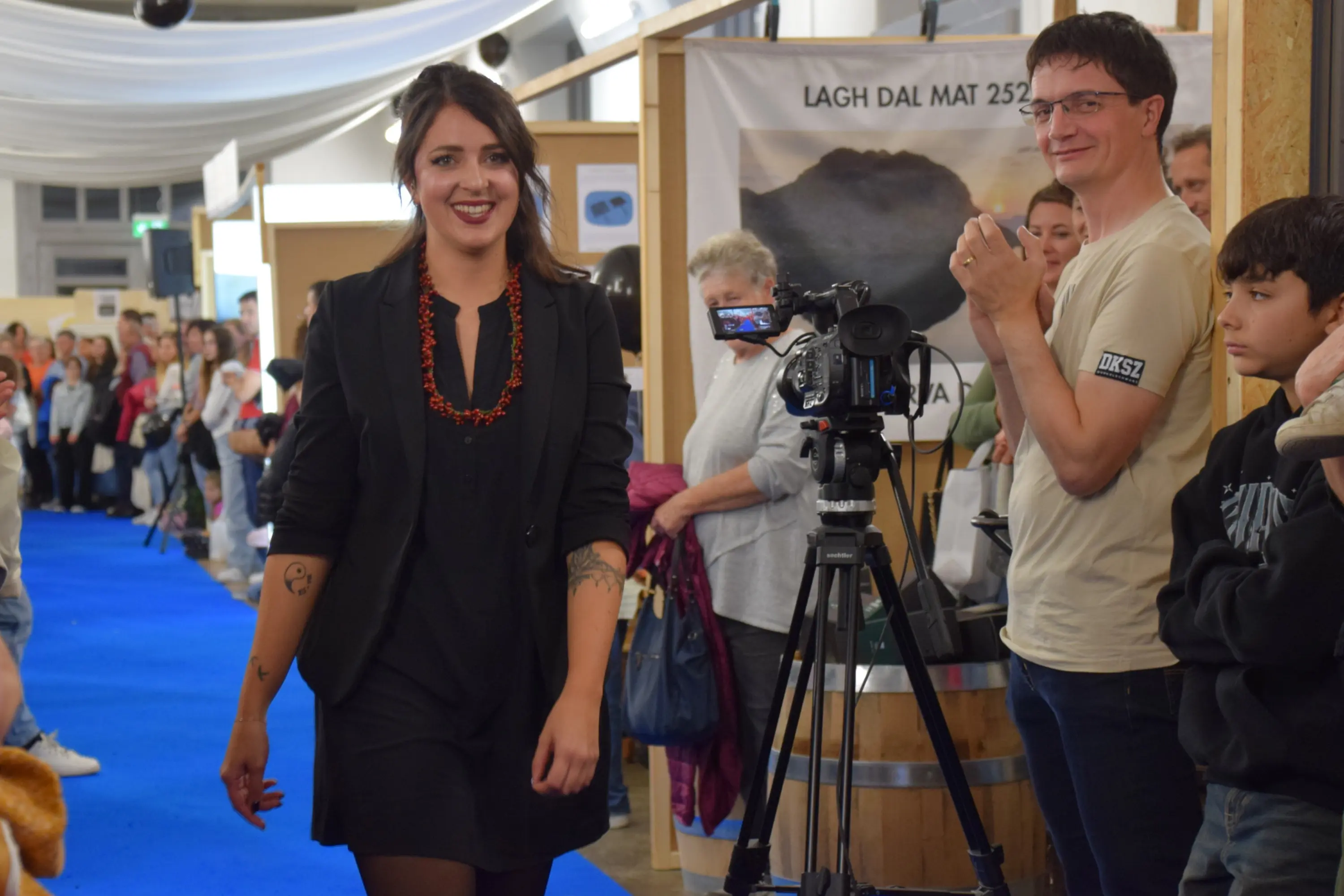 Una donna sorridente con un vestito nero e una collana di perline arancioni cammina su una passerella, mentre un uomo dietro di lei riprende con una videocamera. Intorno, un pubblico interessato osserva l'evento.