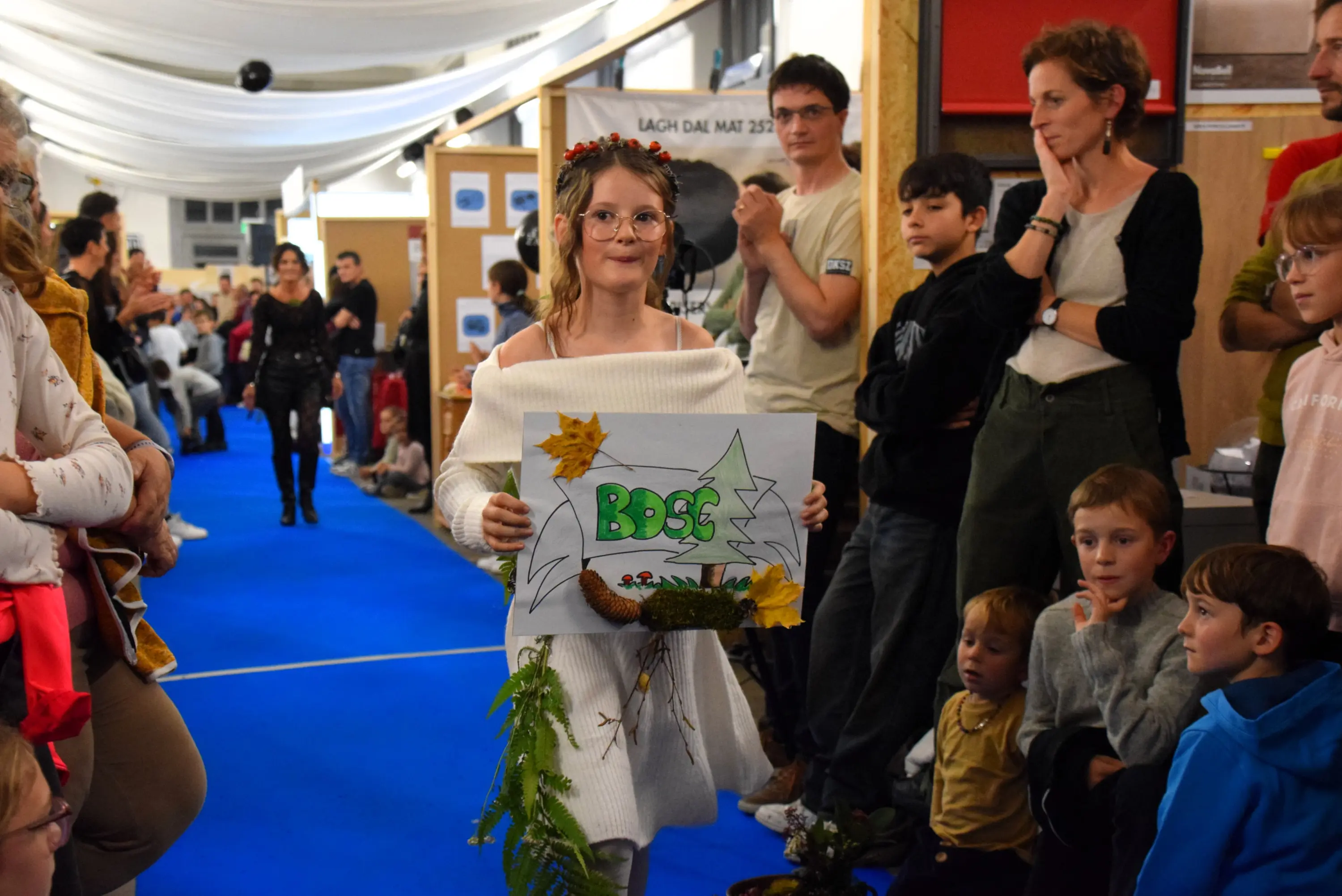 Una ragazza con occhiali e capelli ricci tiene un cartello decorato con foglie e fiori, mentre sfila in passerella. Dietro di lei, un pubblico composto da adulti e bambini osserva attentamente. Il pavimento è di colore blu, e l'amb