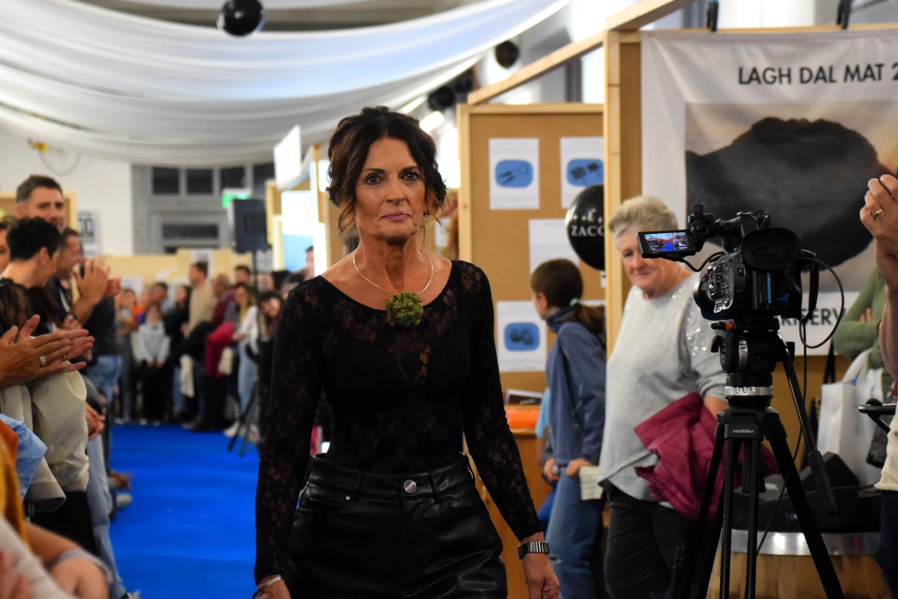 Una modella cammina su una passerella durante un evento, indossando un top nero trasparente e pantaloni in pelle. Il pubblico applaude e sorride intorno a lei. In fondo si vede un cameraman.