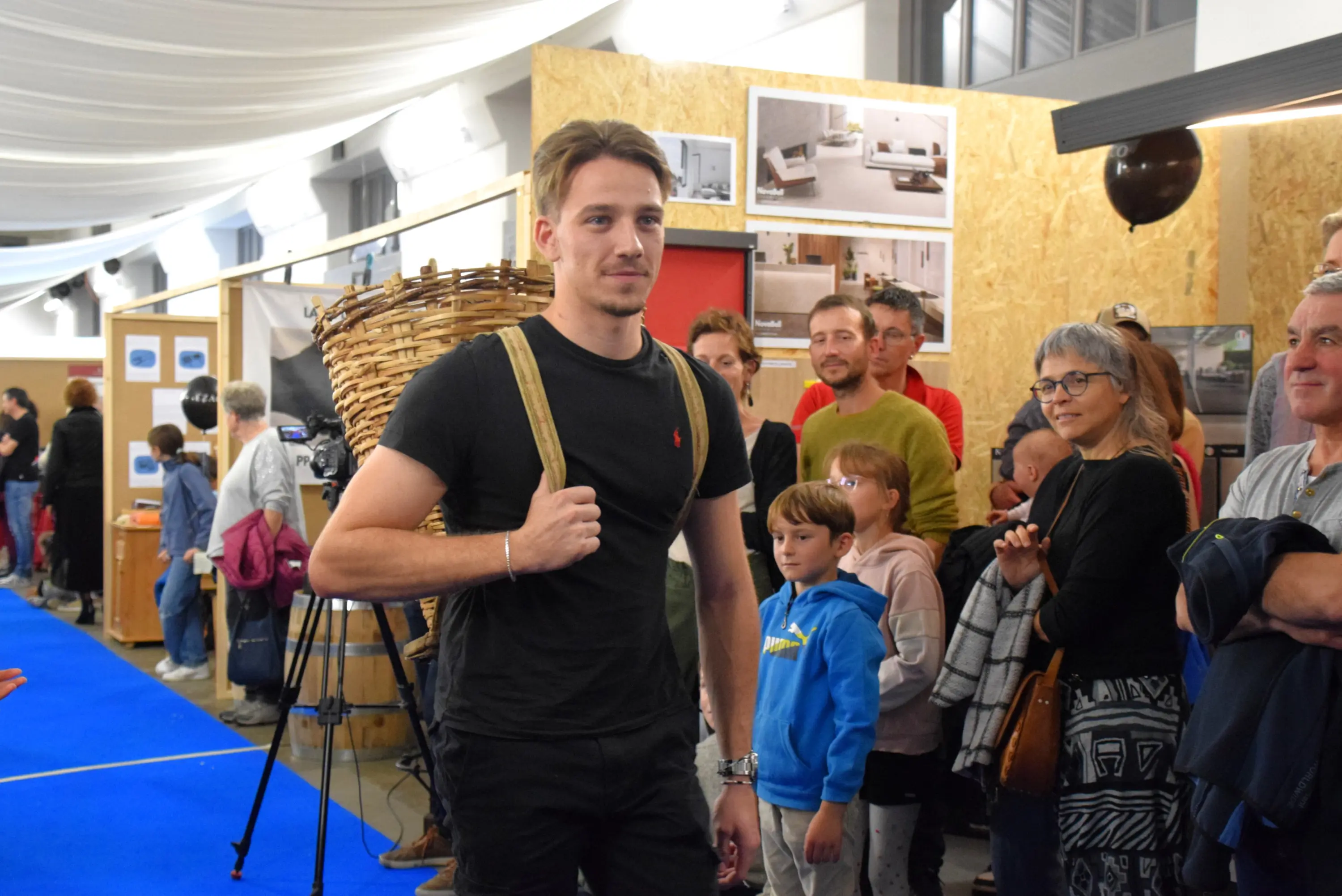 Giovane uomo con una borsa a tracolla in un evento affollato, mentre altre persone lo osservano. Sullo sfondo, stand espositivi con decorazioni in legno.