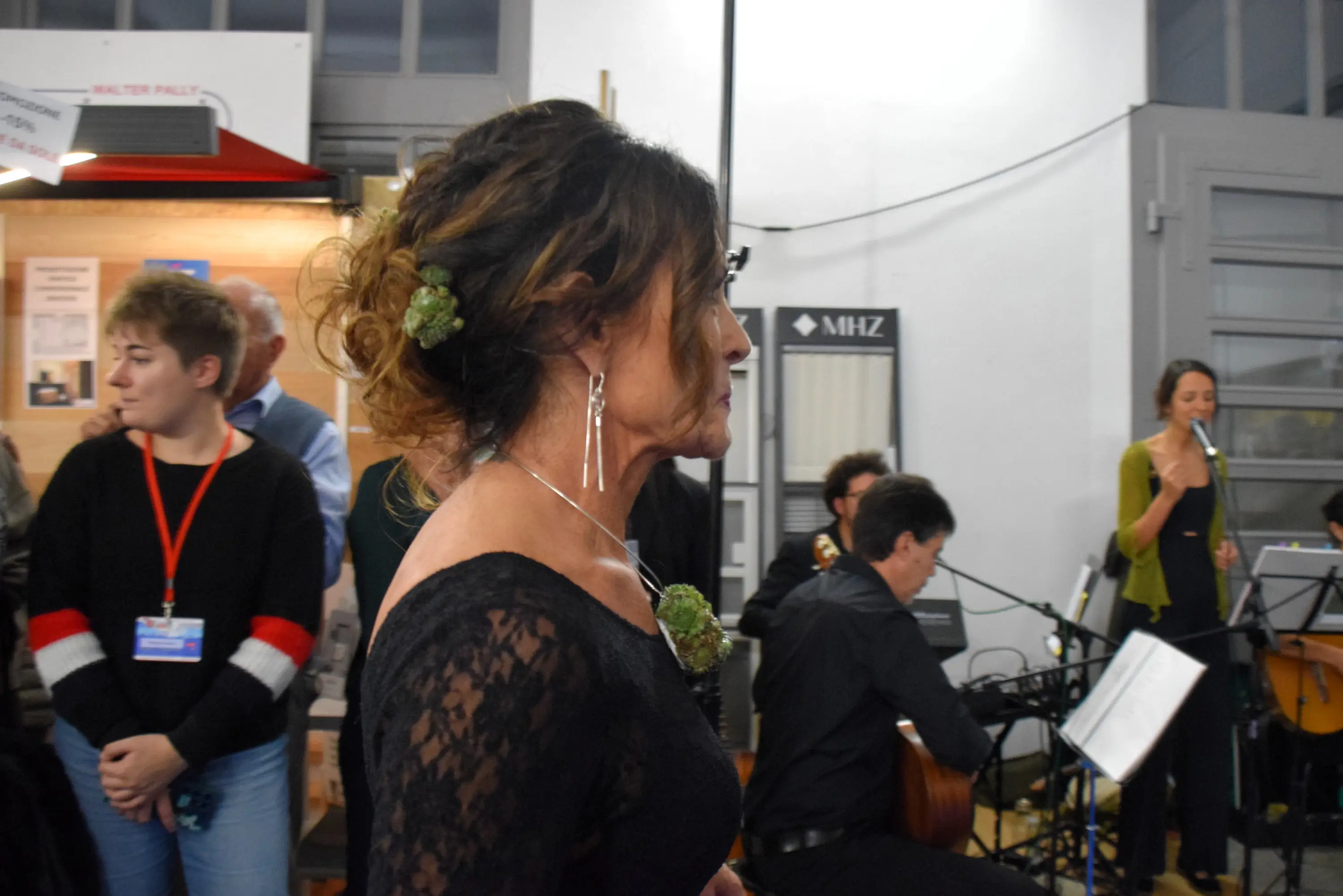 Donna con capelli raccolti e accessorio floreale, indossa un abito nero in pizzo. Sullo sfondo, musicisti suonano strumenti mentre altre persone ascoltano.