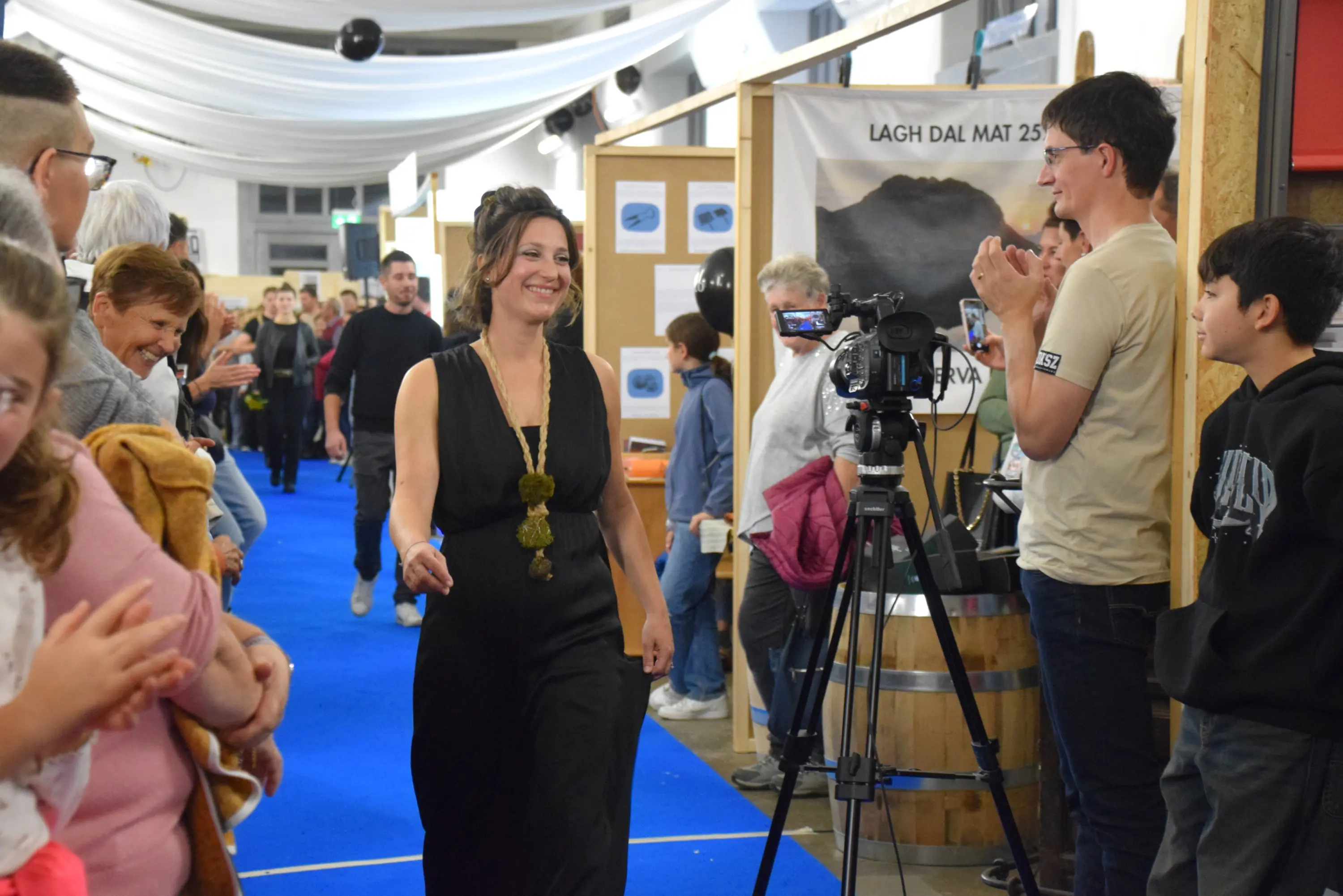 Una donna in abito nero cammina con sicurezza su un tappeto blu mentre il pubblico la applaude. Alcune persone la guardano sorridendo, mentre una telecamera filmato è posizionata davanti a lei. Sullo sfondo, una vista di un evento con decorazioni e