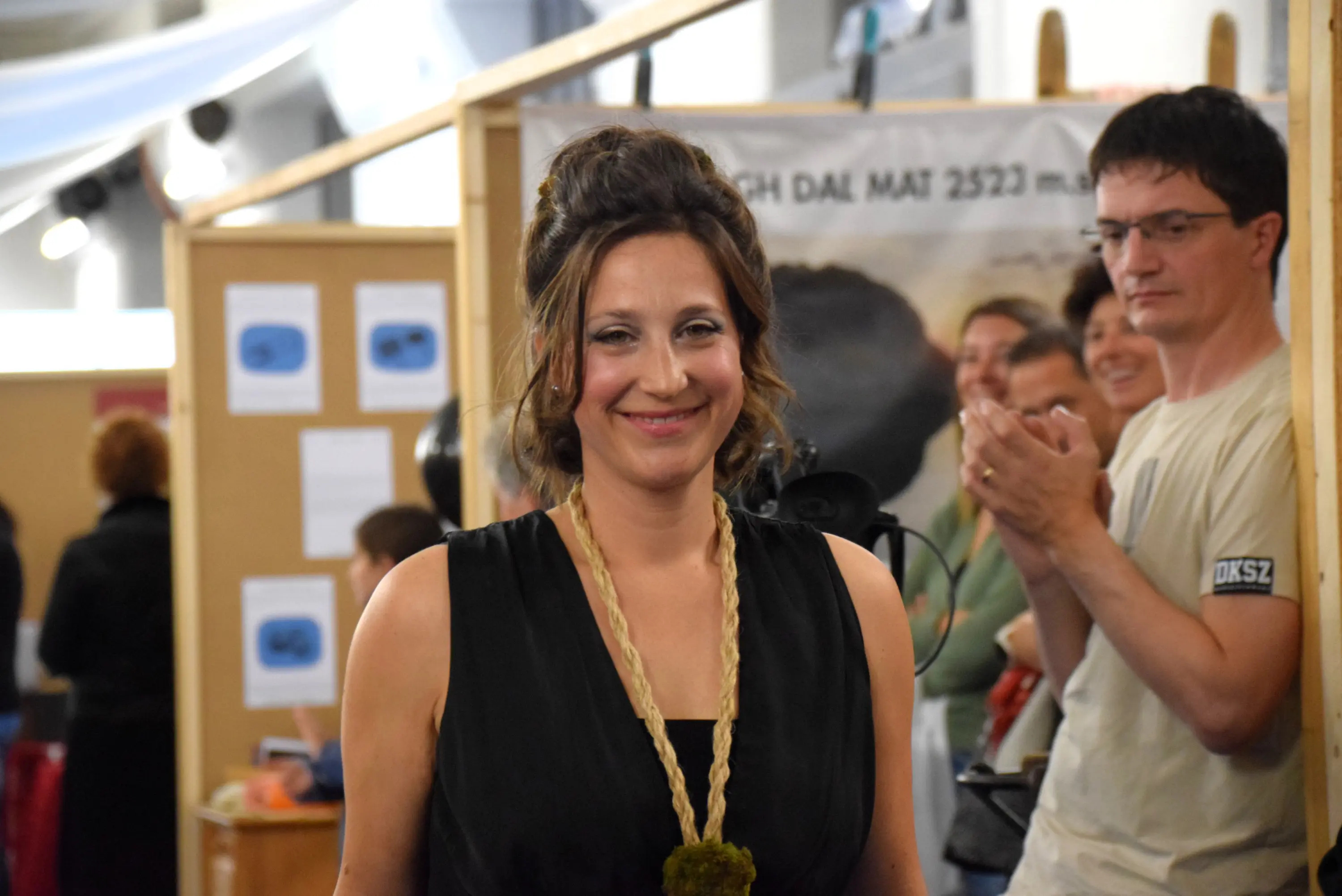 Donna con un abito nero e collana di perline, sorridente mentre cammina verso il pubblico. Sullo sfondo, un gruppo di persone applaude e si nota un ambiente di evento o fiera.