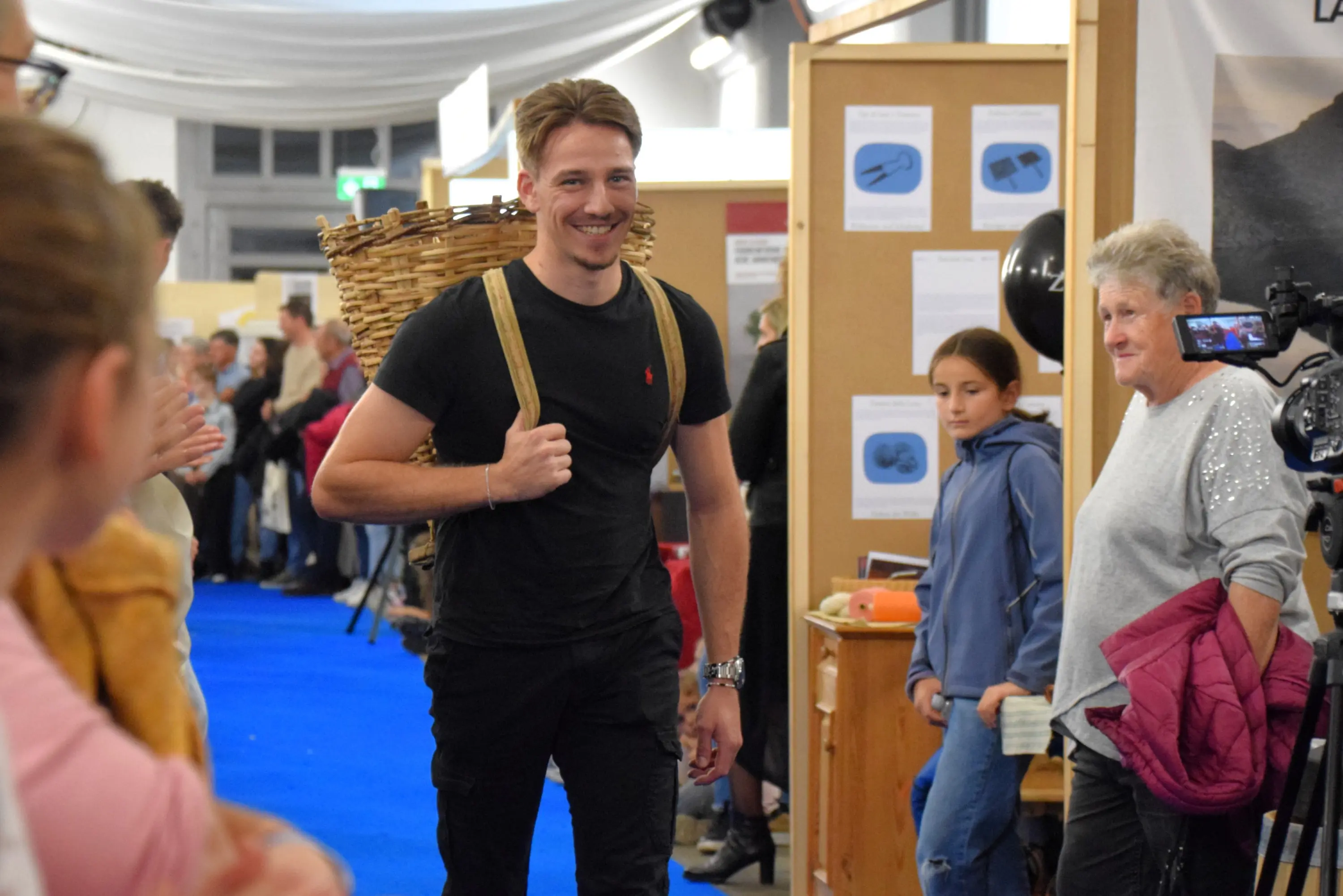 Un giovane uomo sorride mentre cammina con un cesto sulla schiena, circondato da spettatori in un evento fieristico. Due donne lo osservano, una sembra sorpresa. Sullo sfondo, stands e decorazioni dell'evento.