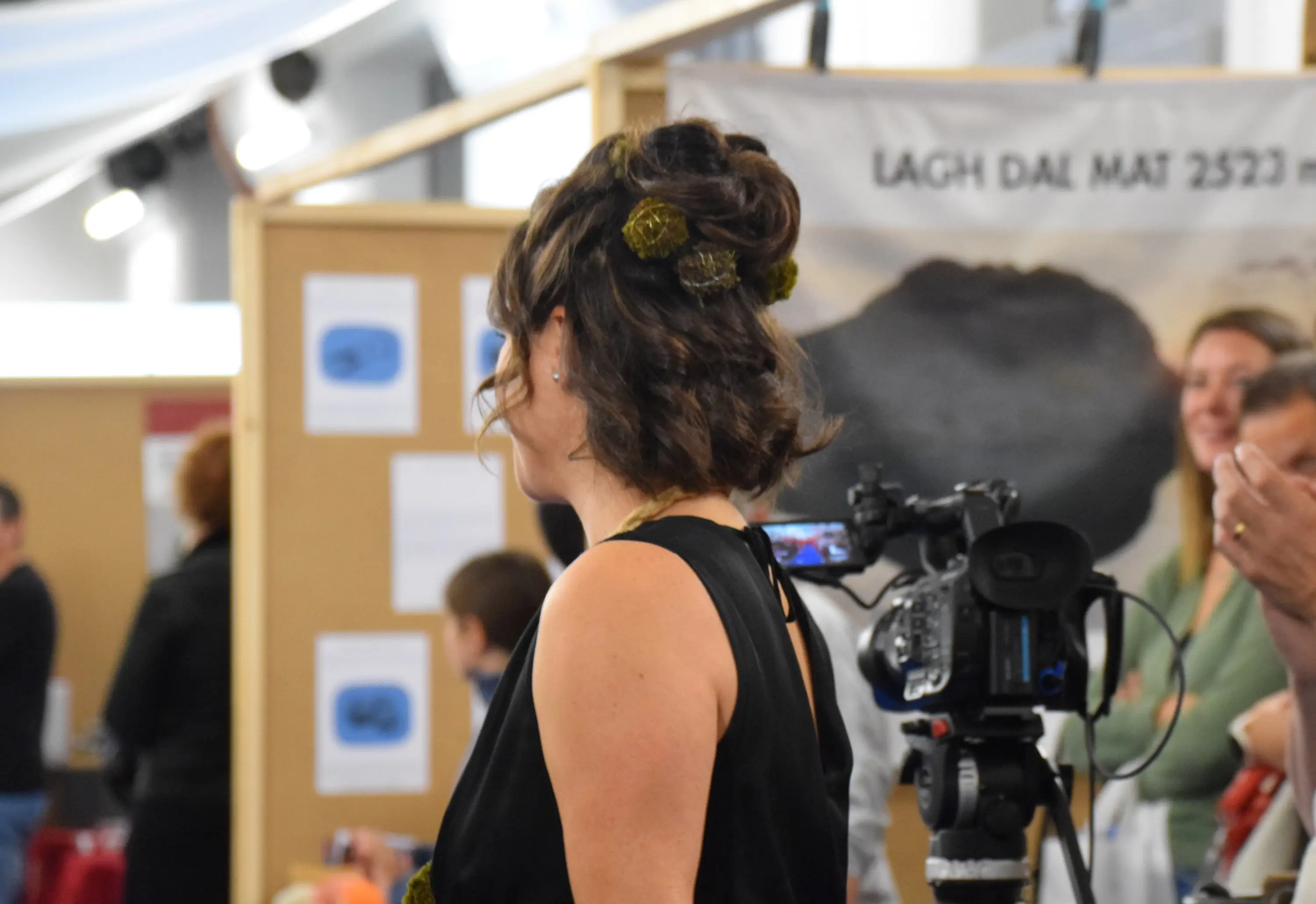 Una donna con capelli ricci e una decorazione floreale osserva un evento. Sullo sfondo, si intravedono alcune persone e attrezzature fotografiche.