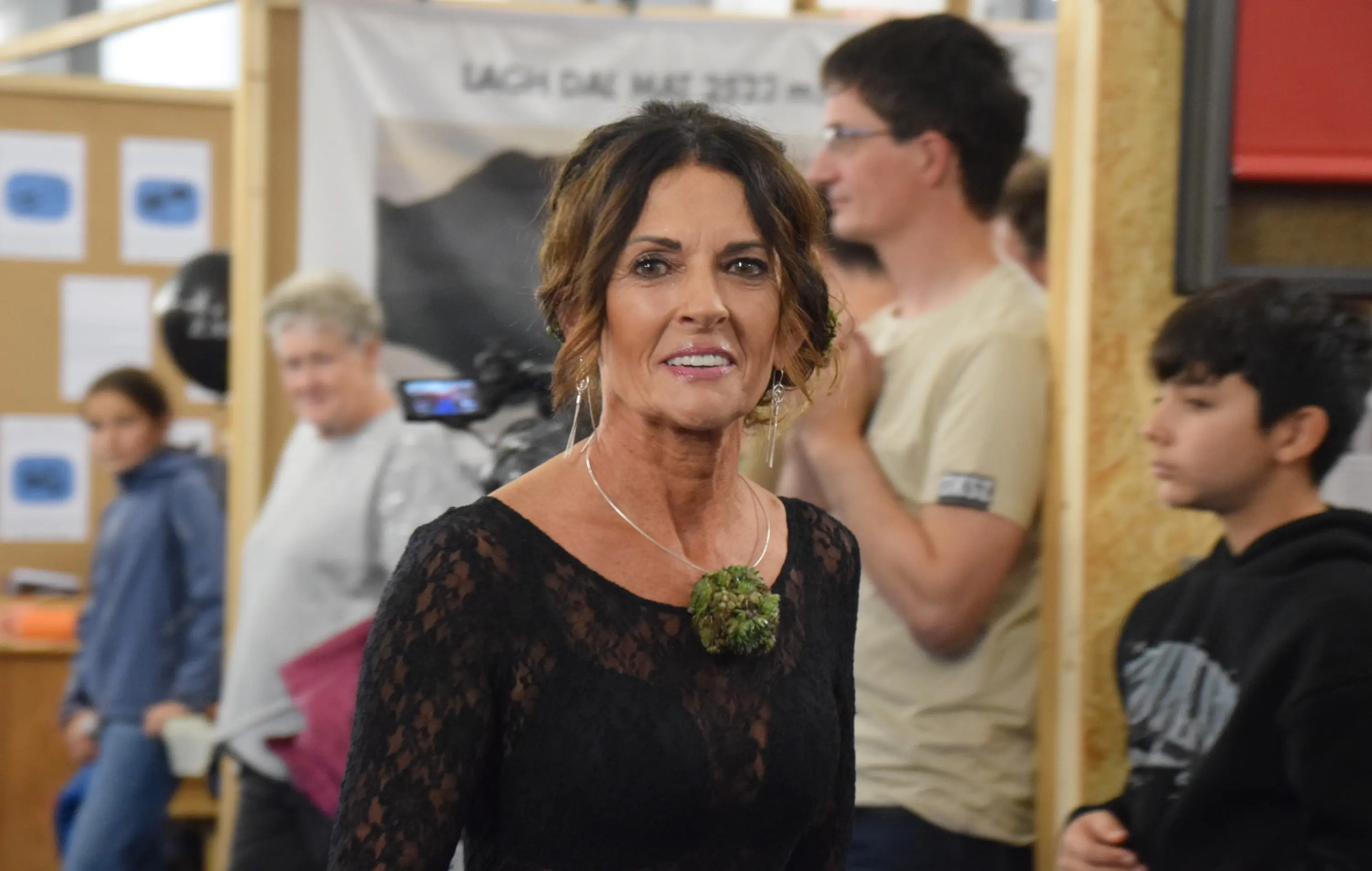 Una donna con capelli scuri e ondulati indossa un abito nero elegante. Ha un collier verde e sorride mentre si trova in un ambiente affollato. Sullo sfondo si vedono altre persone che interagiscono.