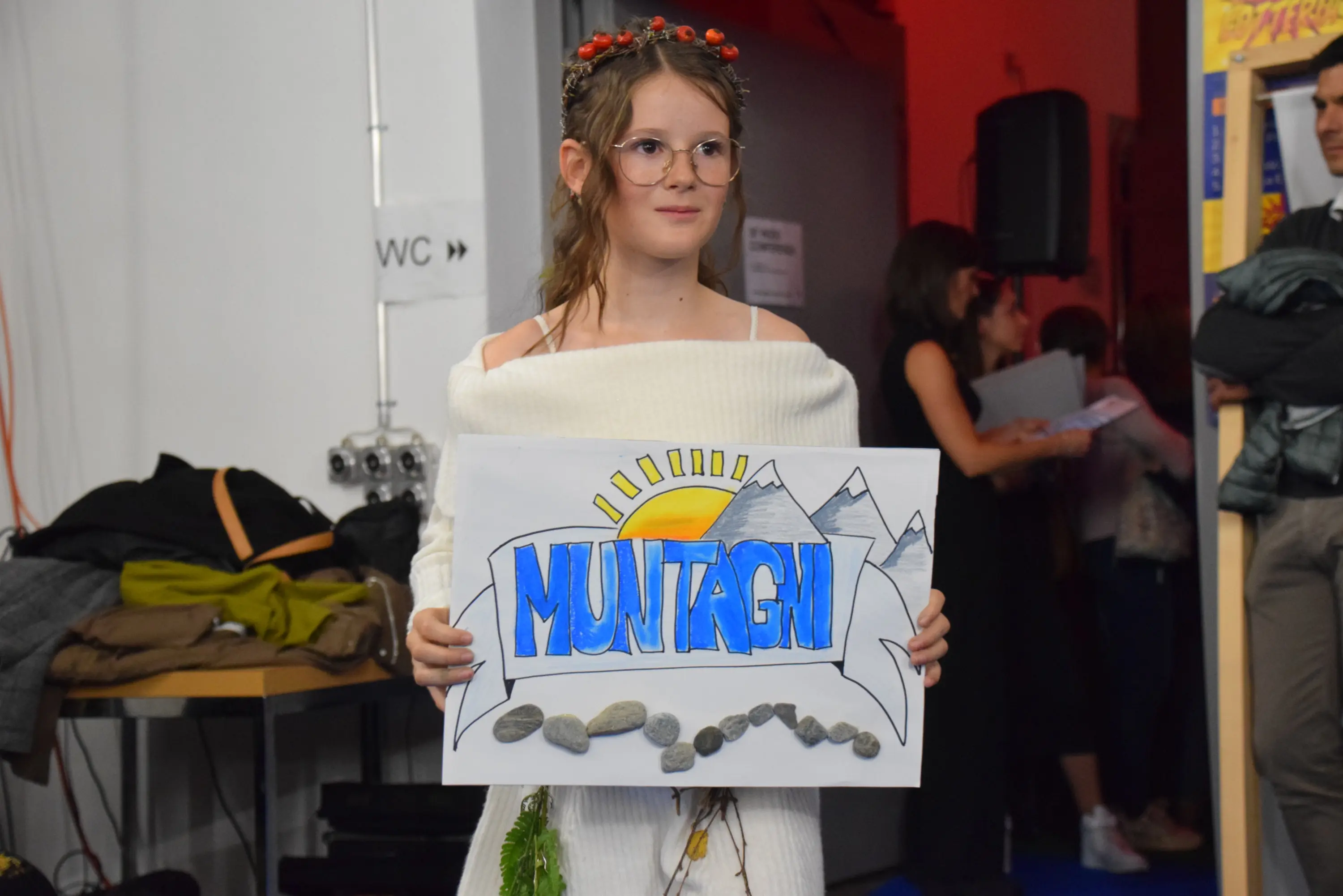 Ragazza con occhiali e abito bianco che tiene un cartello che dice "Muntagn". Ha una corona di fiori e si trova in un ambiente di festa con persone sullo sfondo.