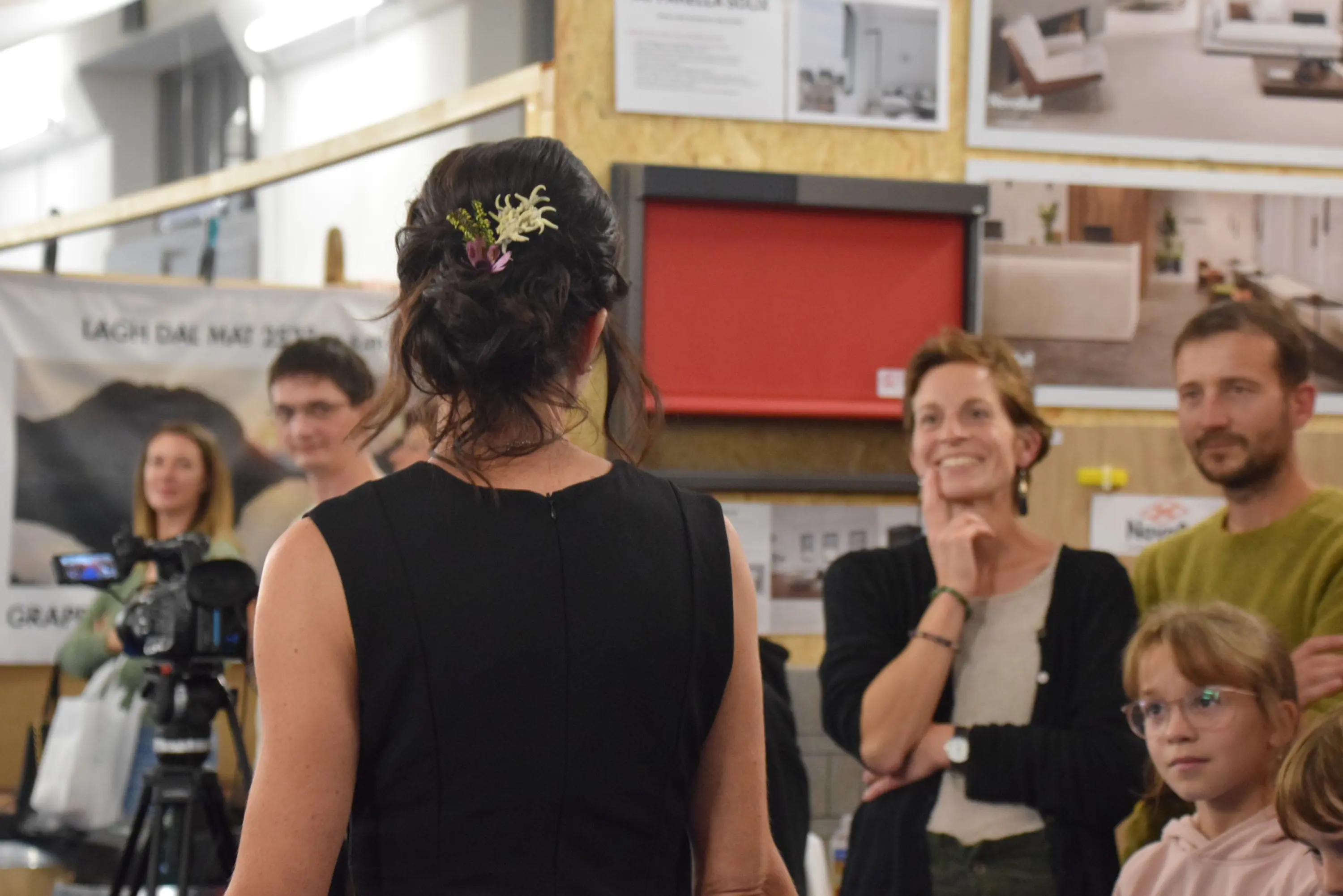 Una donna di spalle con un'acconciatura elegante e un fiore tra i capelli, si trova di fronte a un gruppo di persone sorridenti che la osservano attentamente in un ambiente espositivo.