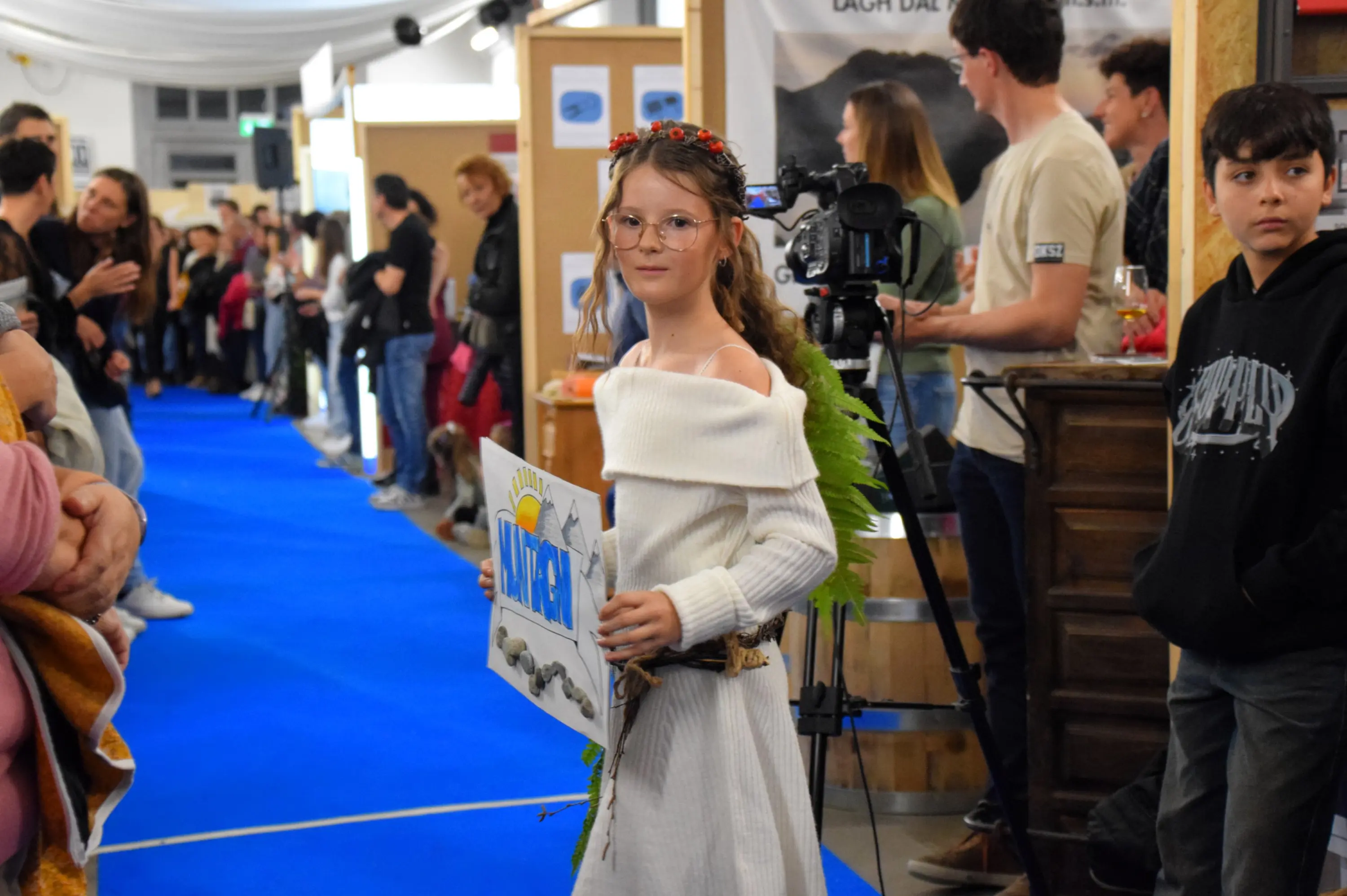Una bambina in abito bianco con decorazioni di foglie tiene un cartello con la scritta "SUN". È circondata da una folla e c'è una telecamera in secondo piano.