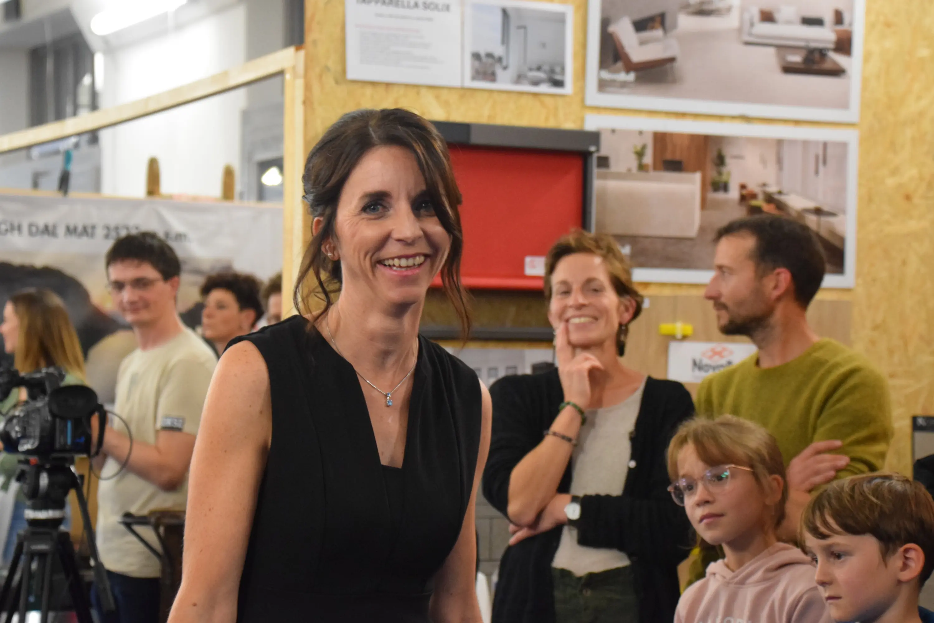 Una donna sorridente in abito nero si trova al centro dell'immagine, circondata da un gruppo di persone, tra cui bambini e adulti, che osservano. Sullo sfondo si intravedono poster e prodotti di arredamento.