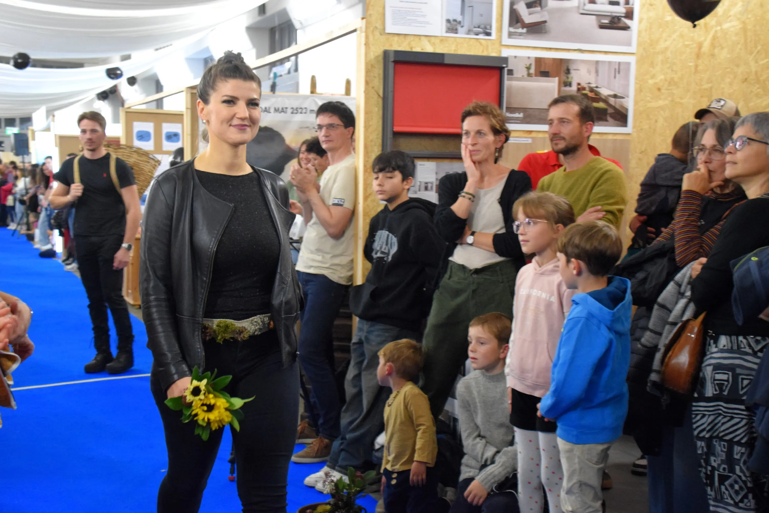 Una donna in abbigliamento nero cammina su un tappeto blu, tenendo in mano un mazzo di girasoli. Intorno a lei, un gruppo di persone di diverse età applaude e osserva, inclusi bambini seduti a terra. La scena si svolge
