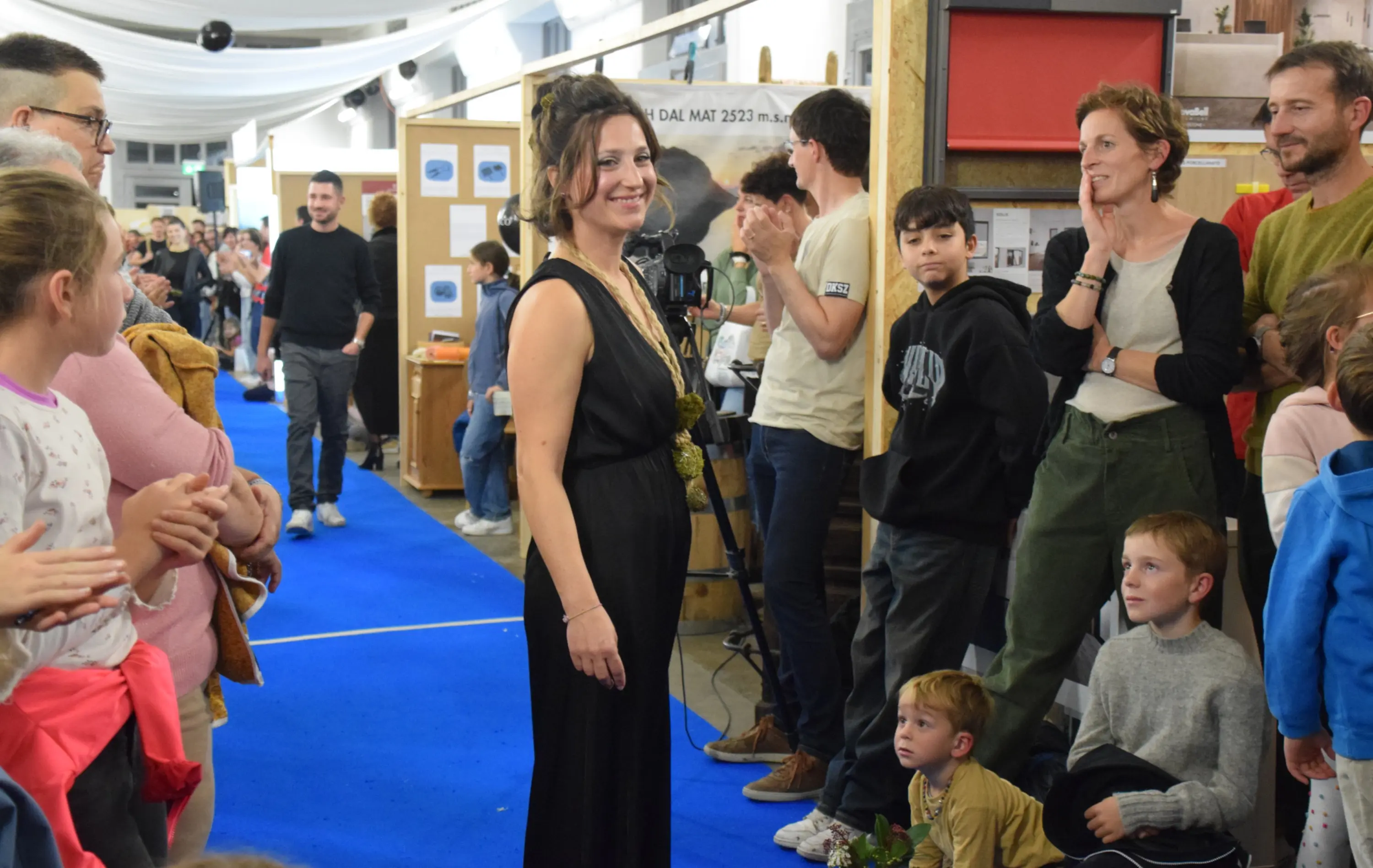 Una donna in abito elegante sorride al centro di una passerella, mentre il pubblico applaude. Intorno a lei, un gruppo di persone, tra cui bambini e adulti, osserva con attenzione. Il pavimento è blu e sullo sfondo si vedono espositori.