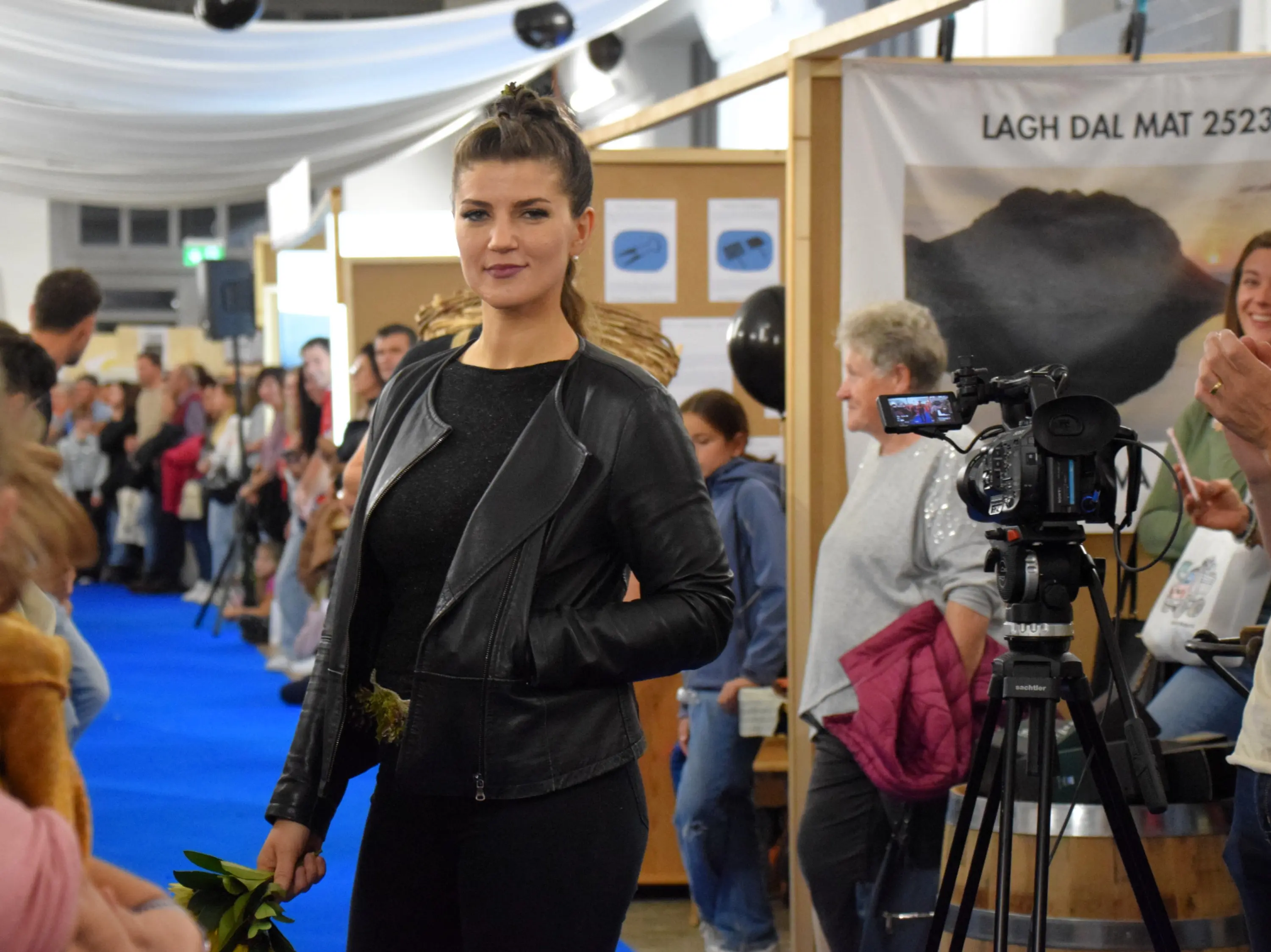 Una modella in passerella indossa una giacca di pelle nera, mentre tiene in mano un mazzo di fiori. Sullo sfondo, si vedono persone che la osservano e una videocamera riprende l'evento.