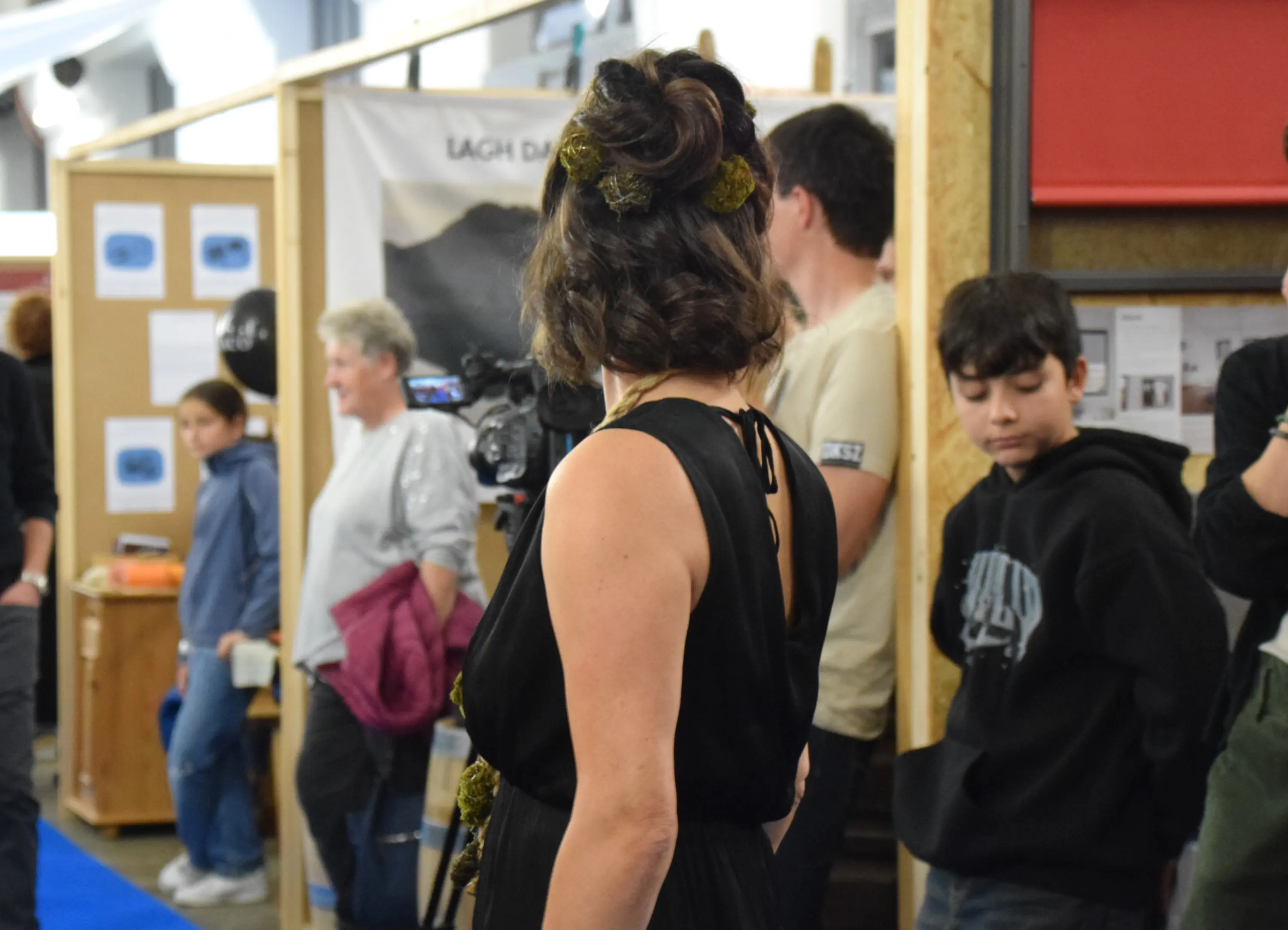 Una donna con capelli ricci e un abito nero osserva il pubblico, mentre alcuni giovani si trovano dietro di lei in un ambiente affollato.