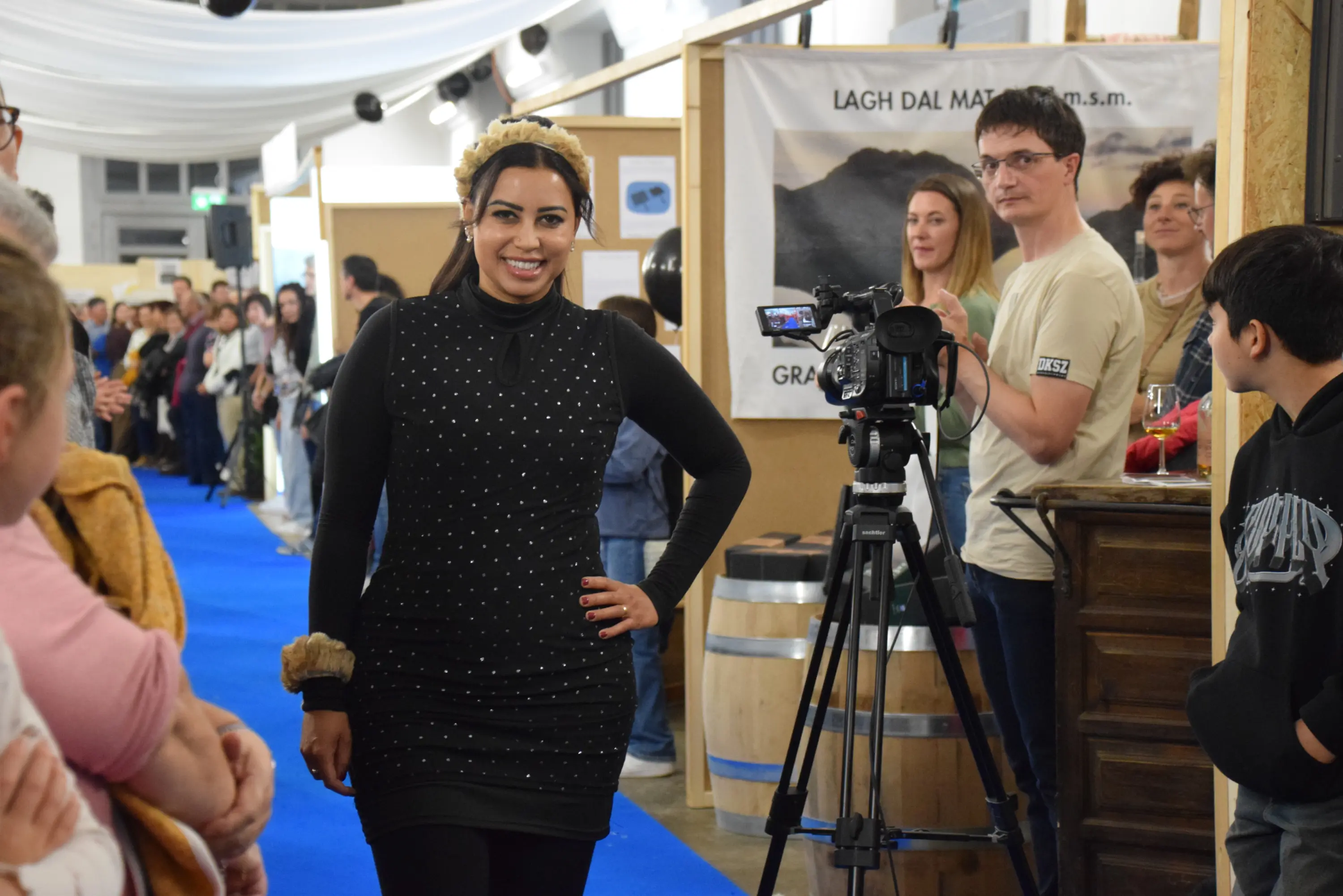 Modella in abito nero con dettagli luccicanti, posando con un sorriso su un tappeto blu. Sullo sfondo, una folla osserva e una videocamera è pronta a riprendere.