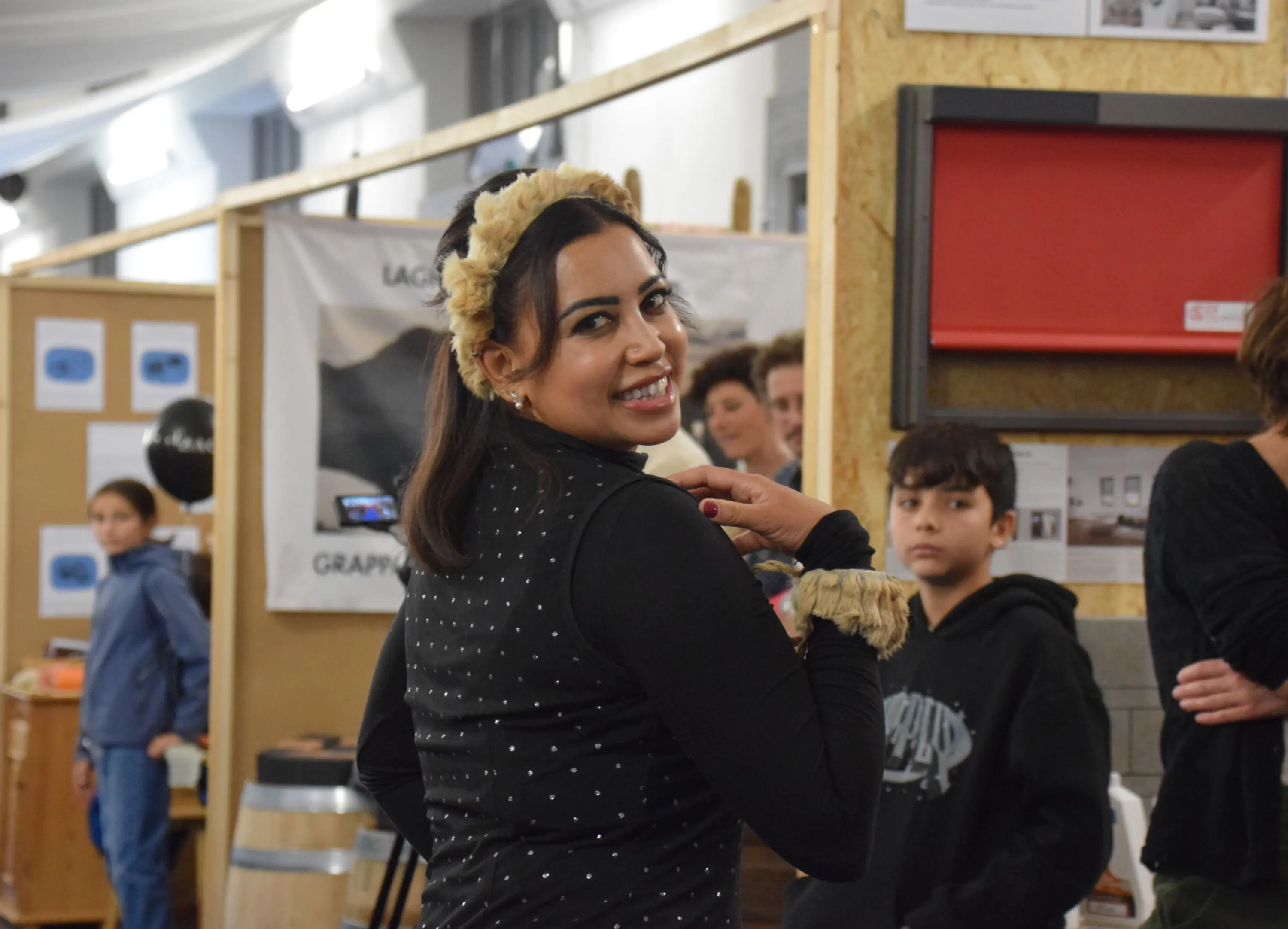 Una giovane donna sorridente indossa un abito nero con decori, mentre si volta verso la telecamera. Ha accessori decorativi sui capelli e si trova in un ambiente espositivo con persone e materiali visibili sullo sfondo.