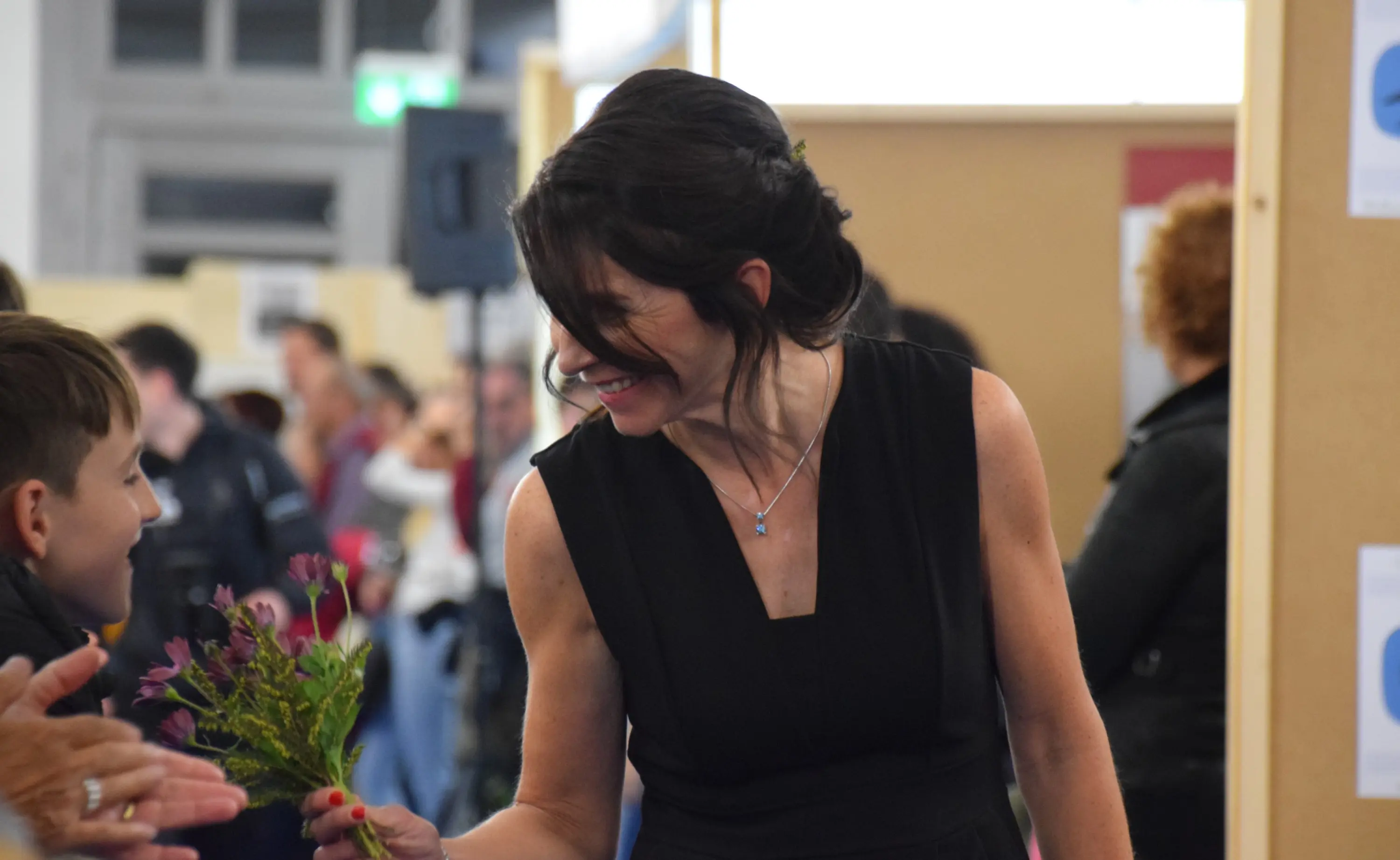 Una donna con abito nero riceve un mazzo di fiori da un ragazzo, sorridendo. Sullo sfondo, si intravedono altre persone e un ambiente affollato.