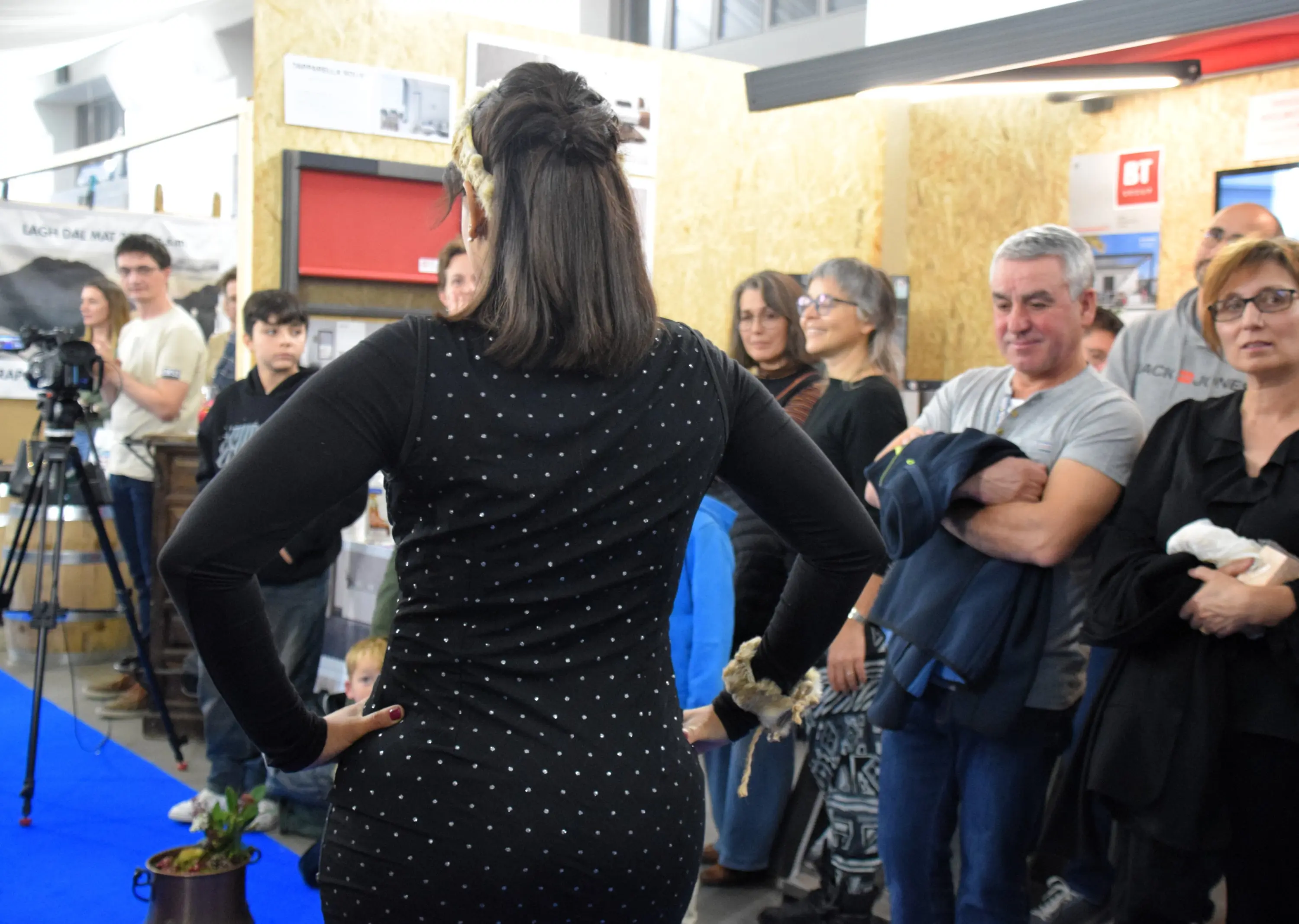Una modella di spalle inizia una sfilata, indossando un vestito a pois. Un pubblico di varie età osserva con interesse, alcuni in piedi e altri seduti, in un ambiente vivace con espositori sullo sfondo.