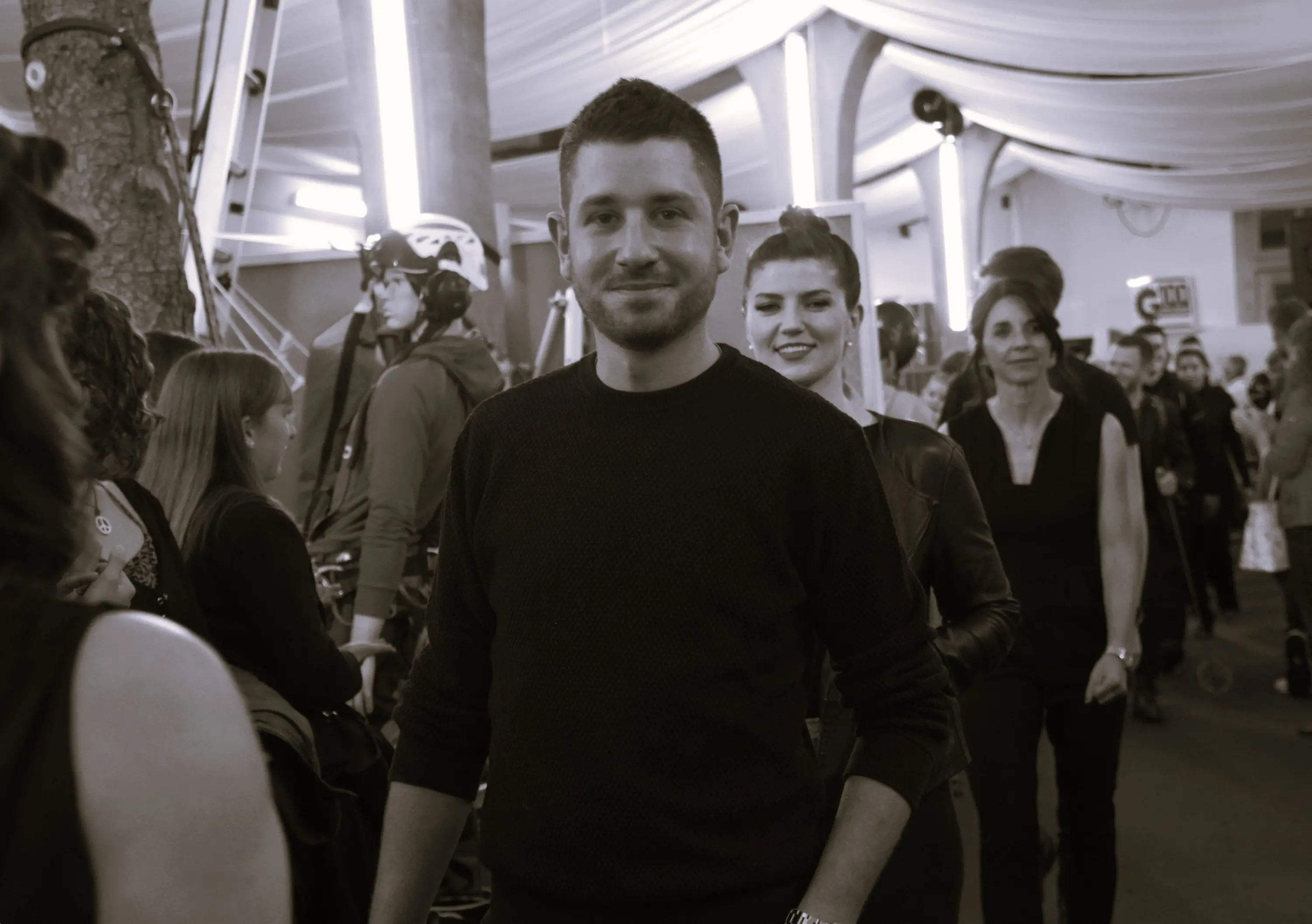 Un uomo sorridente in maglione nero si trova in un ambiente affollato, con diverse persone che lo seguono. Sullo sfondo, si possono vedere attrezzature e decorazioni.