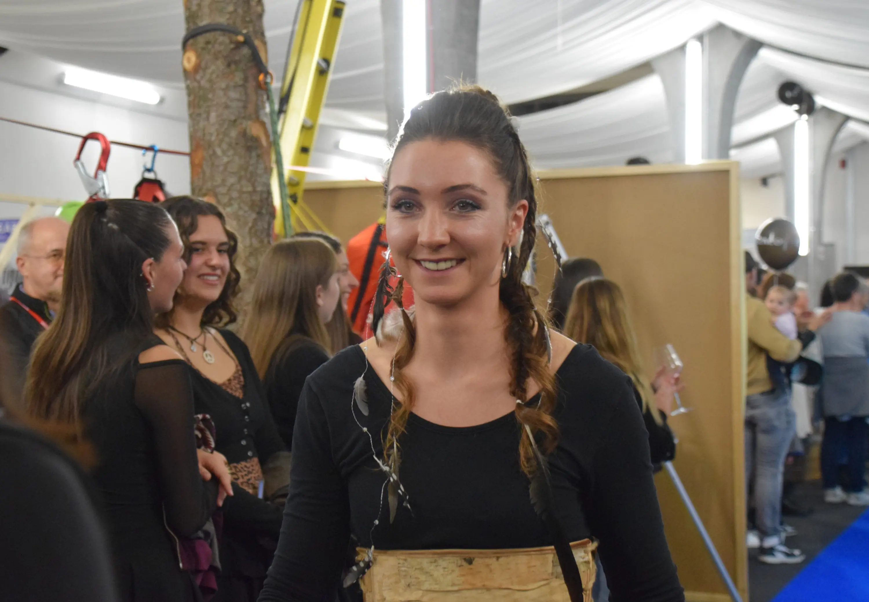 Una donna sorridente con capelli lunghi e trecce, vestita in abbigliamento nero, si trova in un ambiente festoso con altre persone sullo sfondo.