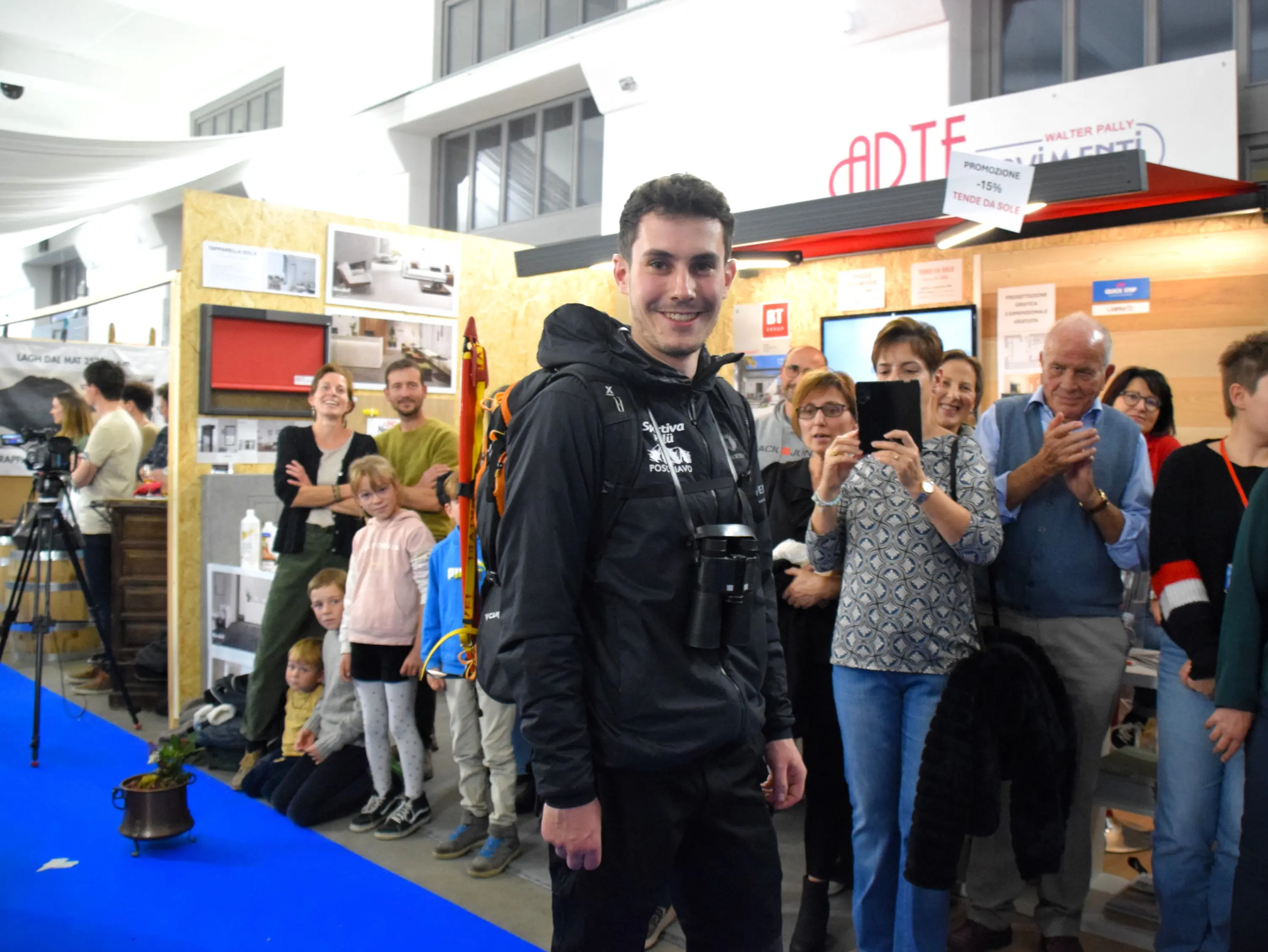 Un giovane con uno zaino e una macchina fotografica sorride in primo piano, mentre dietro di lui un gruppo di persone, tra cui famiglie e adulti, lo osserva con interesse in un evento o una fiera.