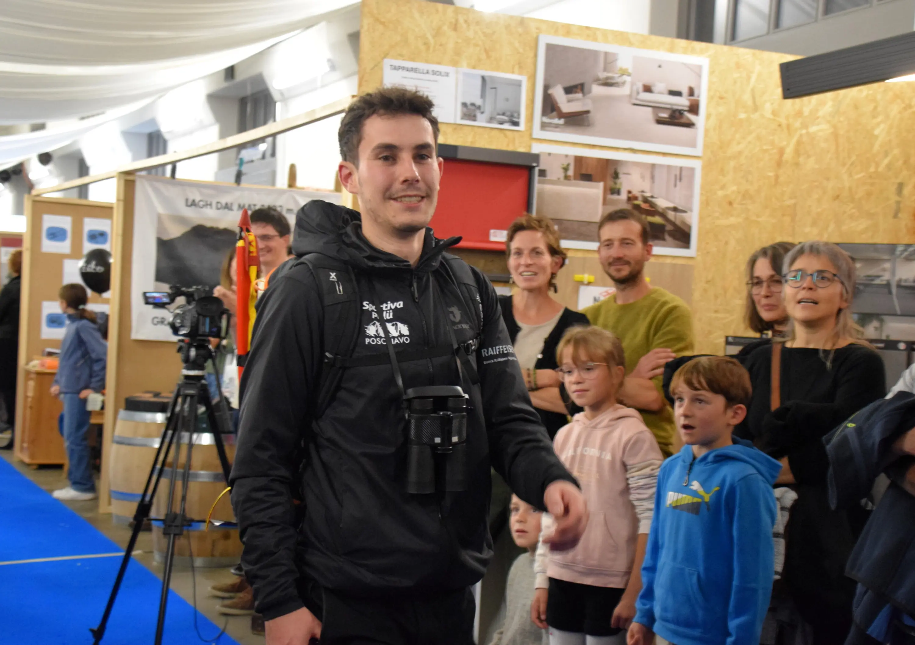 Un giovane con una fotocamera al collo sorride mentre si dirige verso la folla in un evento. Sullo sfondo, diverse persone, tra cui bambini e adulti, osservano interessati.
