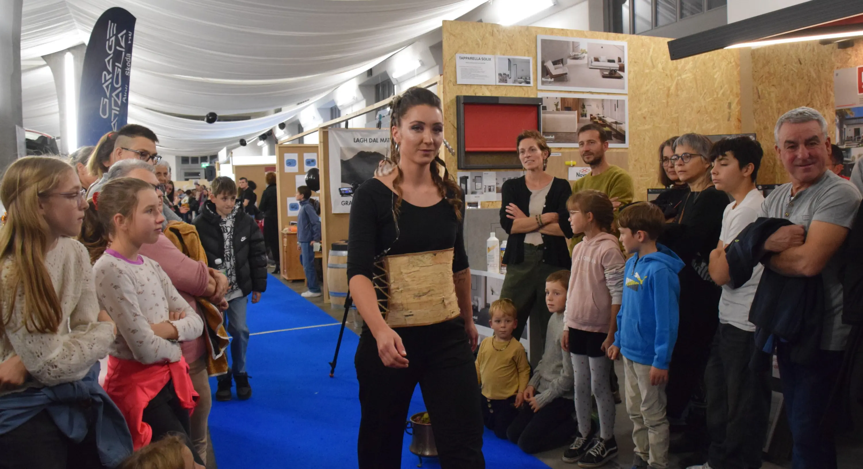 Una modella presenta un outfit alla fiera, circondata da un pubblico composto da adulti e bambini, tutti attenti e interessati. La passerella è decorata con un tappeto azzurro, mentre sullo sfondo si vedono espositori.