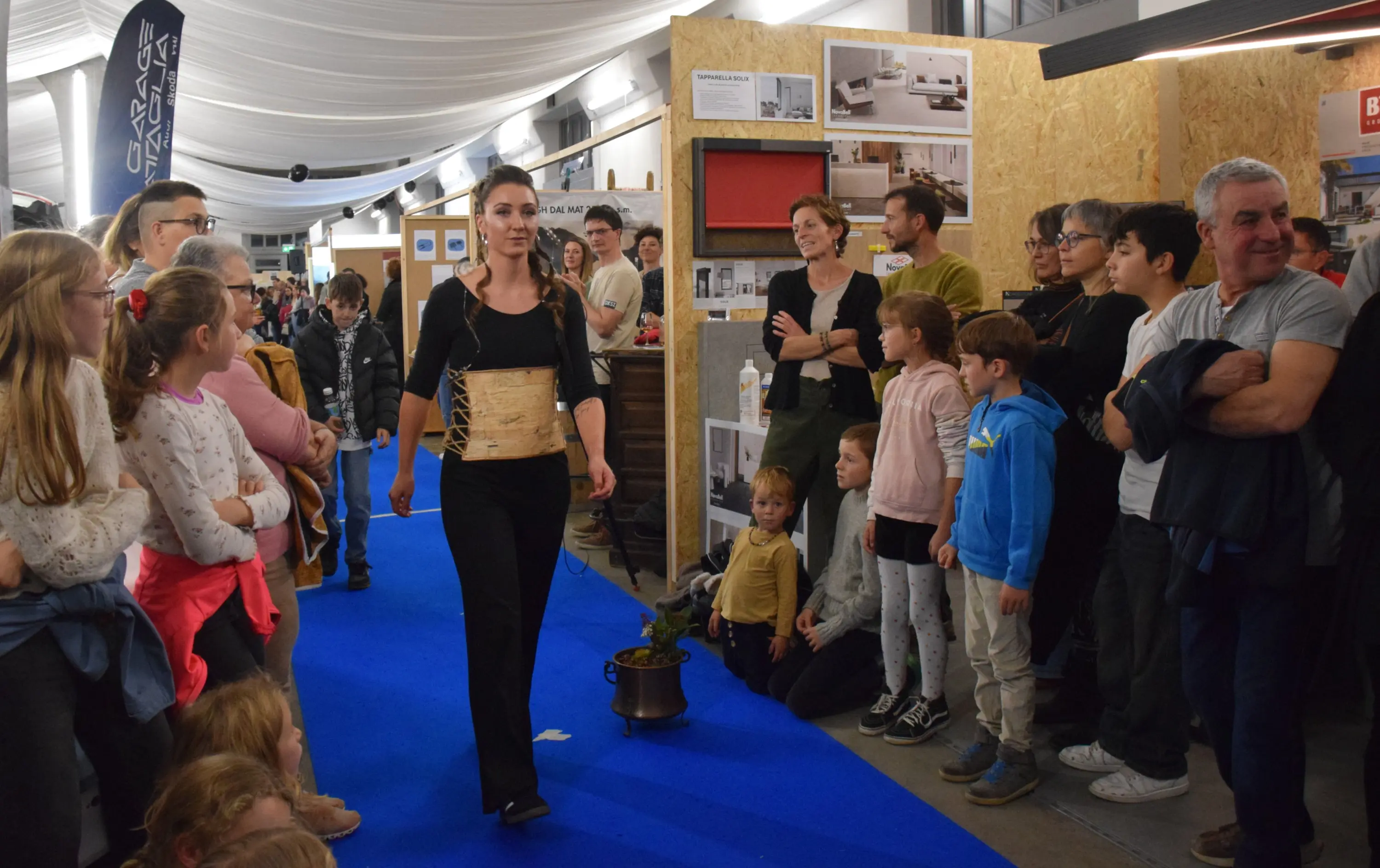 Una donna in abbigliamento raffinato cammina su un tappeto blu mentre un gruppo di persone, tra cui bambini e adulti, la osserva attentamente. Alcuni partecipanti appaiono interessati e curiosi. Sullo sfondo si intravedono esposizioni e altri